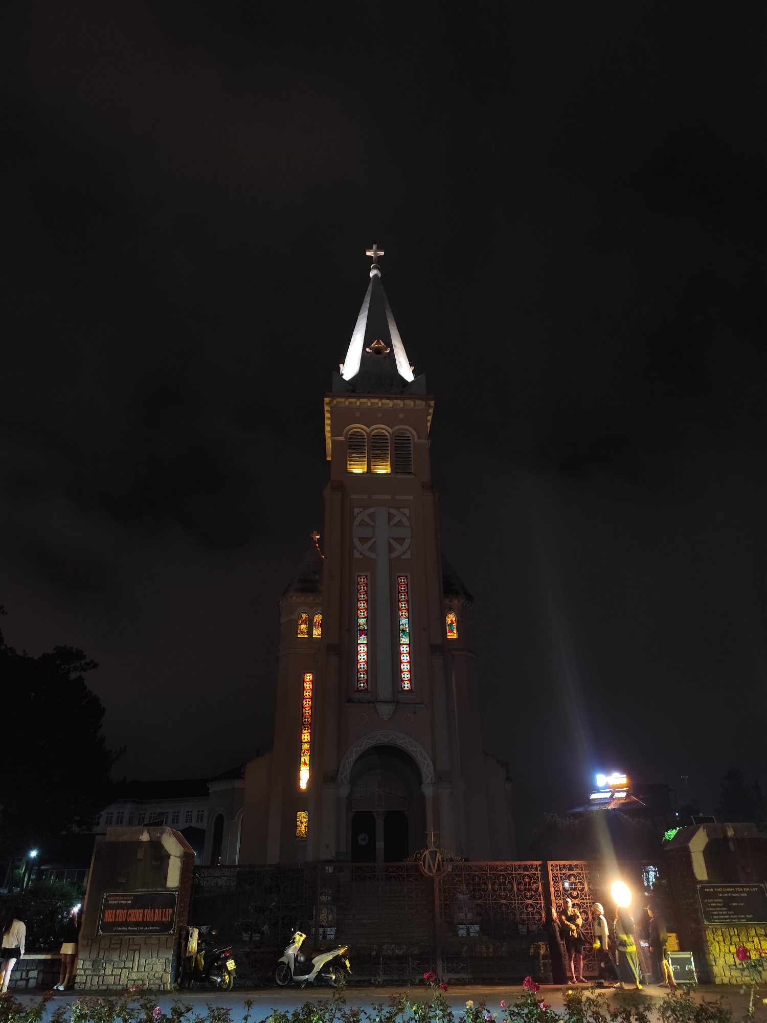 At night, the Domaine de Marie Cathedral in Da Lat, Vietnam, stands tall against a dark sky. The cathedral's steeple is illuminated, topped with a cross. Stained glass windows line the walls. A low wrought-iron fence and some signs in front of the cathedral are visible. A group of people stand near a bright light source, possibly setting up equipment. Parked motorbikes are situated near a stone sign. The scene is framed by flowers and low vegetation.