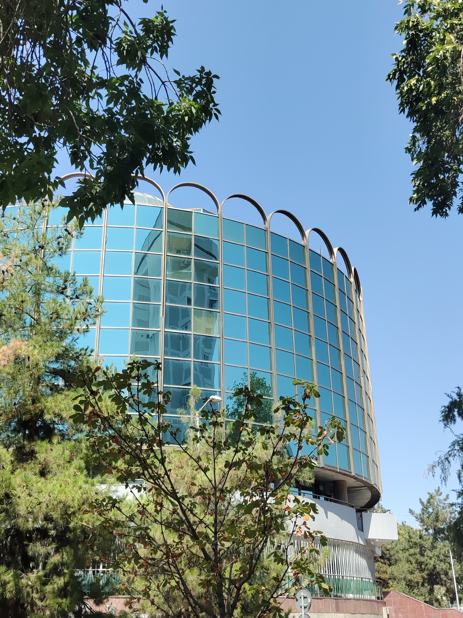 This image shows a modern, curved building with a facade of blue glass windows and decorative arched metal elements at the top. The building reflects the sky and a portion of another building, which appears to be part of a symmetrical structure. The building is surrounded by lush green trees, some with cones, and other foliage. The sky is a clear, bright blue. The image is taken from a low angle, looking upwards towards the building. The building's design suggests it is an office building, hotel, or a similar public or commercial structure. The architectural style is contemporary and sleek. The setting appears to be urban, with the presence of trees suggesting a park or a well-landscaped area within a city. The building's curvature and reflective glass are prominent features.
The building in the image is the International Business Center, also known as Tashkent City Business Center, located in Tashkent, Uzbekistan. It is a prominent landmark in the city's modern skyline.