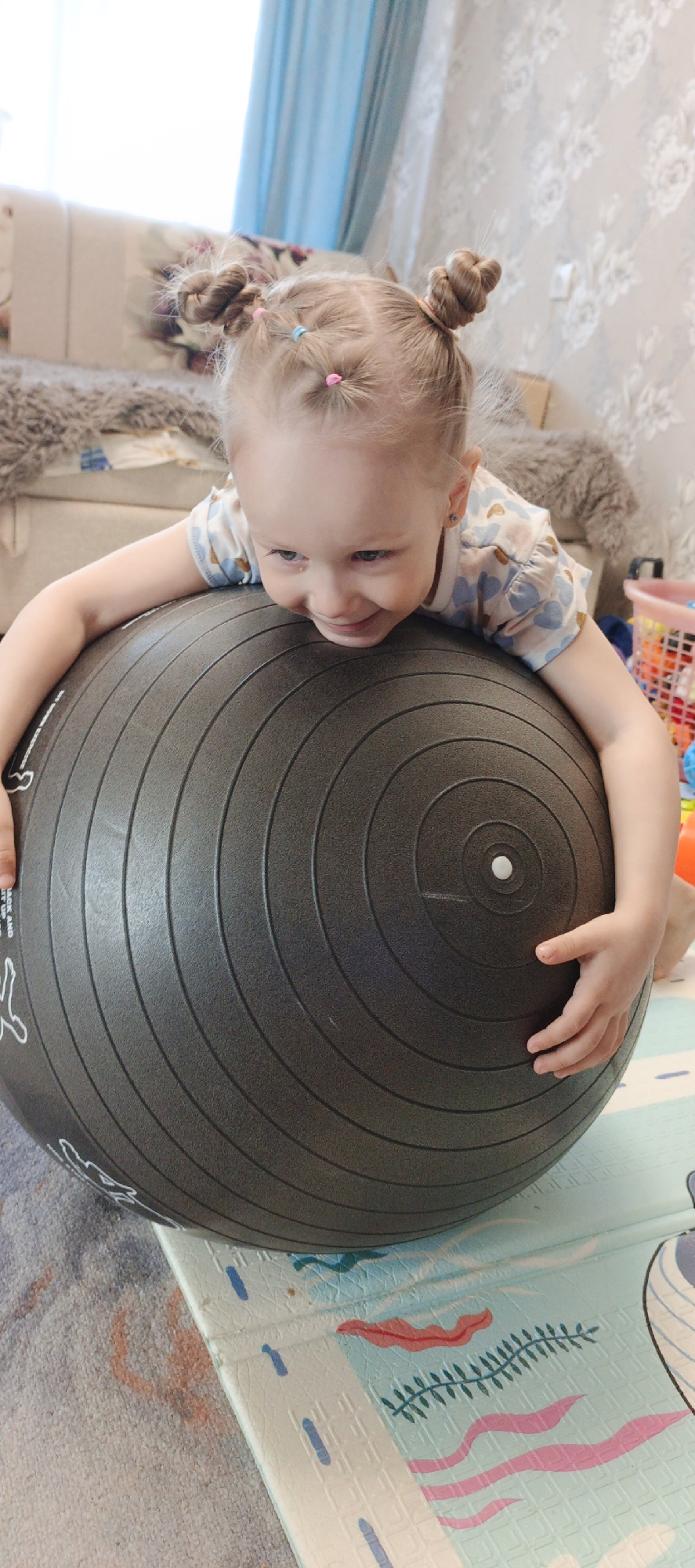 A young, fair-skinned child, likely a girl, lies on her stomach across a large, dark grey exercise ball, gripping it with both hands. Her blonde hair is styled in multiple small bun-like pigtails, secured with pink and light blue elastic bands. She has a wide, happy smile on her face, with a playful expression, and her blue eyes are looking downwards. She is wearing a short-sleeved top with a light background and a pattern of light blue and peach-colored hearts or cloud-like shapes. Small earrings are visible in her ears.
The exercise ball, the central object, is dark grey with concentric circular grooves and a white valve cap. White outline drawings of figures performing exercises, along with some largely illegible white text, are visible on its surface.
The setting is indoors, in what appears to be a child's room or play area in Chaikovsky, Russia. In the blurred background to the left, a light-colored sofa or bed with patterned pillows and a textured grey blanket can be seen. To the right, a light-colored wall with subtle floral or leaf-patterned wallpaper is visible. A light blue curtain is in the upper left corner. The floor beneath the ball consists of a patterned play mat featuring light blue, pink, and green marine-themed designs (like waves, seaweed, and stylized fish), partially covering a light-colored carpet. A pink plastic basket filled with colorful toys is partially visible on the far right.
The child appears to be actively engaged in play or exercise, demonstrating joy and excitement. The lighting suggests it is daytime.