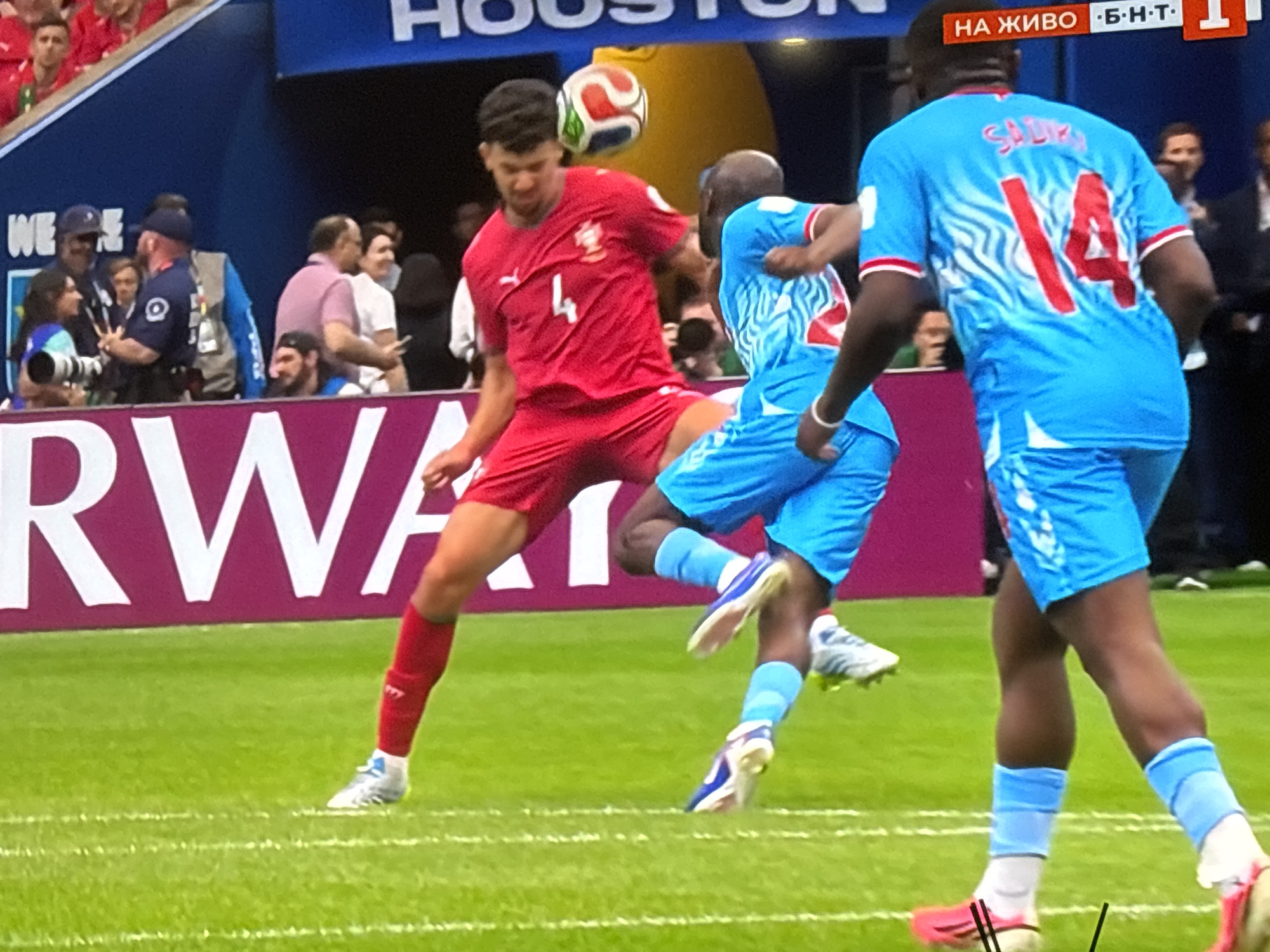This media file captures a dynamic moment during a soccer match. Three male soccer players are the main subjects, engaged in active play on a vibrant green grass field. One player in a red jersey, marked with the number '4', is in mid-air, attempting to head a soccer ball that is positioned just above his head. His body is contorted in effort, with his legs bent and his gaze focused on the ball.
Opposing him, two players in blue jerseys are also involved in the play. One, wearing the number '14' and the name "SALIB" on his back, is positioned behind the player in red, his arms raised as if anticipating a header or attempting to block. The other player in blue is to the right, also in motion, with his legs positioned for play. The soccer ball itself is a prominent object, multi-colored with red, white, and green sections, captured in sharp focus.
The scene is set within a stadium, indicated by the blurred background which shows spectators in the stands, some wearing red. A large banner or advertisement with bold white lettering on a dark background is partially visible behind the players. A digital scoreboard displaying Cyrillic text and numbers is also visible in the upper right corner. The setting appears to be outdoors, and the lighting suggests it's daytime. The overall atmosphere is one of intense athletic competition.
Visible text includes "НА ЖИВО Б•Н•Т 1" on a red and orange banner in the upper right, likely indicating "Live" and a channel or broadcast name. Another partial text appears on the dark banner in the background, potentially a sponsor or team name. The implied emotion is one of concentration and athletic exertion. The location is contextualized as Burgas, Bulgaria.