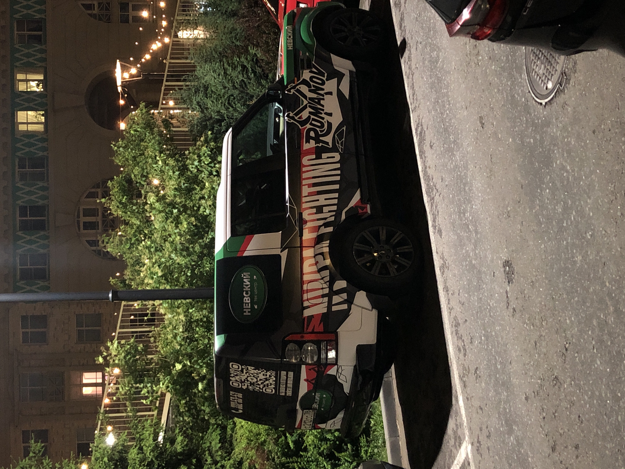 A black Land Rover Discovery 4 SUV is parked on an asphalt surface at night. The vehicle is adorned with a custom wrap featuring colorful graphics, including the text "FIGHTING ROMANOV" in large, stylized lettering and a QR code. The rear of the vehicle displays a license plate that reads "c212cc 78". Behind the SUV is a building with illuminated windows and a metal railing with string lights, surrounded by lush green trees. The scene appears to be located in Saint Petersburg, Russia, indicated by the building's architecture and the license plate format. No people are visible in the image.