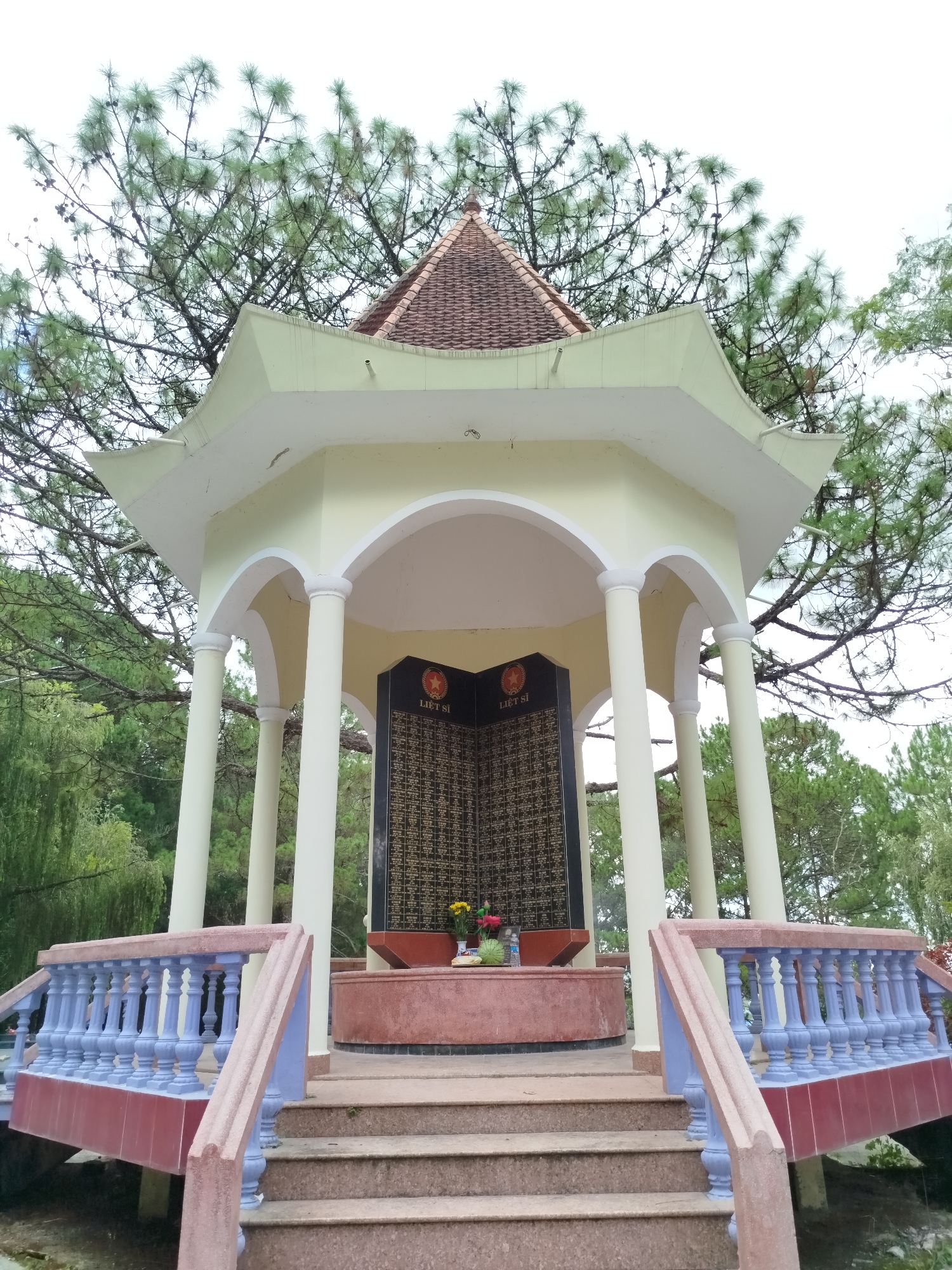 The image depicts a memorial structure located in the city of Cam Ly - Đà Lạt, Vietnam. The structure is a small, ornate pavilion with a tiled, peaked roof, supported by white columns. Inside the pavilion is a dark plaque inscribed with names and adorned with two red stars. Steps lead up to the pavilion, and blue and maroon railings surround the base. The pavilion is situated outdoors, surrounded by trees.