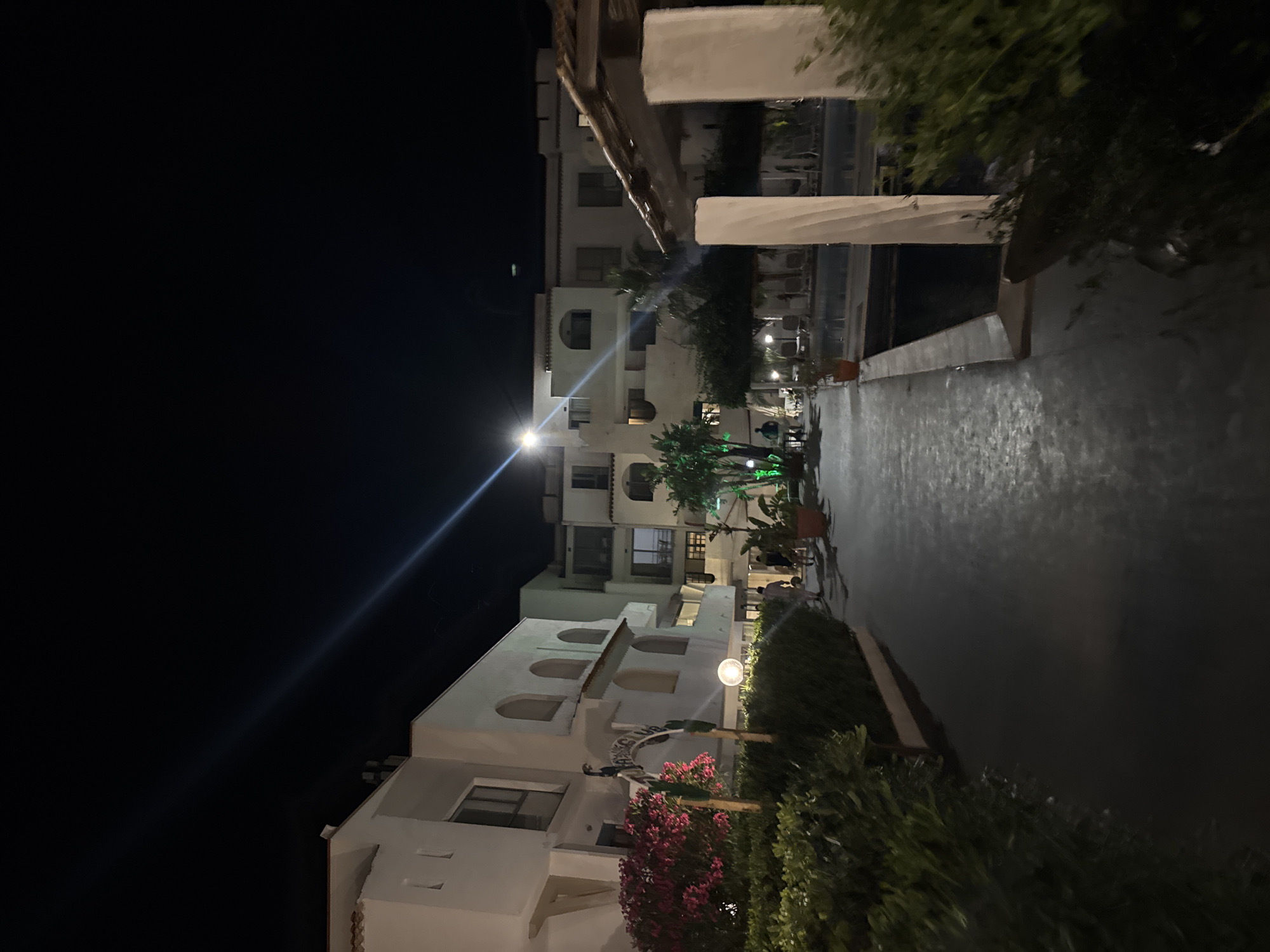 The image depicts a nighttime view of a resort or hotel complex in Isola delle Femmine, Italy. In the foreground, a paved walkway runs between lush green bushes and hedges on the left and a swimming pool area on the right. Several white pillars, partially obscured by foliage, line the edge of the pool. The buildings, characterized by their white facades and arched windows, are illuminated, casting a soft glow on the surroundings. A bright beam of light, likely from a spotlight, cuts across the dark night sky. The presence of lounge chairs and a few scattered potted plants suggests a leisure setting. The scene is quiet, with a few indistinct figures walking in the mid-ground, indicating normal activity at the resort.
