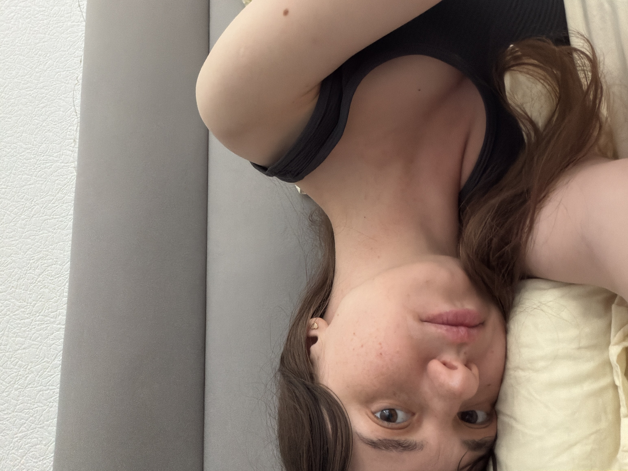 An overhead shot captures a young woman with brown hair, lying on a pillow in bed. Her face is turned towards the camera, with freckles visible on her cheeks and nose. She is wearing a dark-colored tank top. The background is a light-colored textured wall. The scene is indoors and appears to be a bedroom.