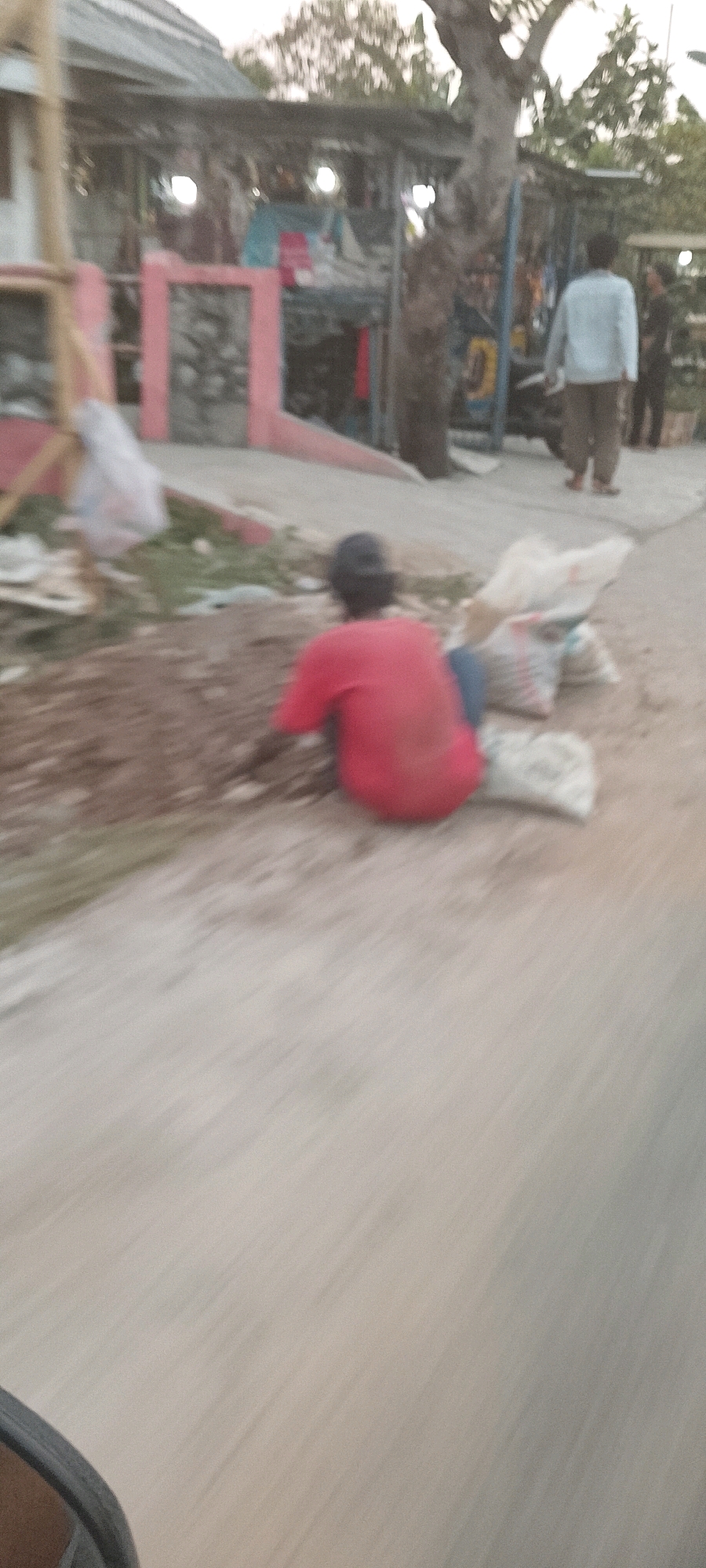 In Sukamaju, Indonesia, a person wearing a red shirt and a dark cap sits on the ground with bags of sand next to them. The person is facing away from the camera, and appears to be working or resting on a dirt path. In the background, a building with a tin roof and what appears to be a market stall are visible. Several other individuals are present in the background, some walking and others standing. The scene is blurred due to motion, suggesting the photo was taken from a moving vehicle.