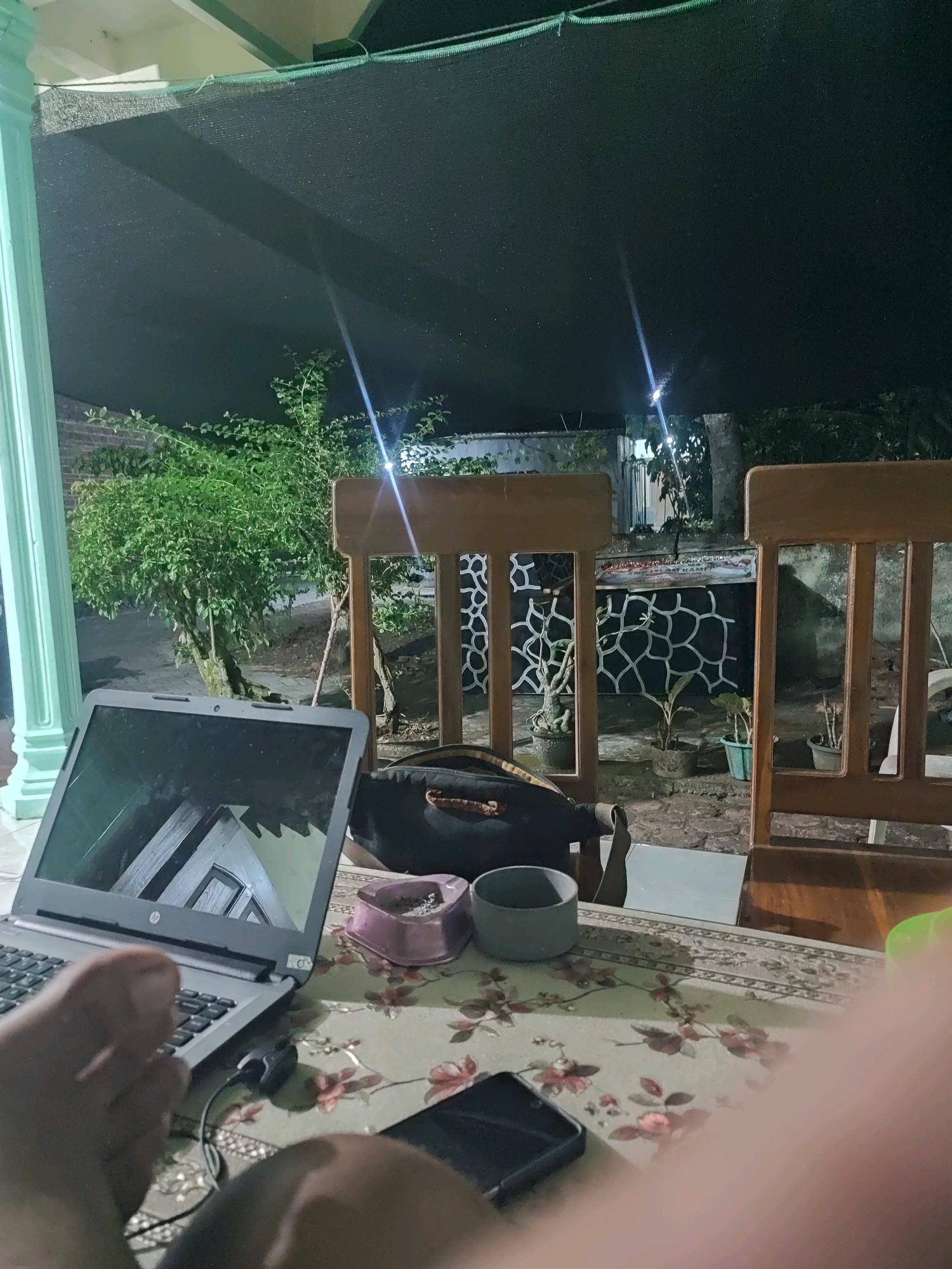 An overhead, low-light, outdoor shot features a table set with a laptop, a phone, two cups, and a pink ash tray. The table is covered with a floral-patterned cloth. In the background, two wooden chairs face a wall with a decorative pattern. A tree and potted plants are visible. The scene is illuminated by a light source above and to the left, casting shadows and reflections. The laptop screen displays a reflection of an indoor environment. The overall setting suggests a domestic outdoor area, potentially a patio or porch.