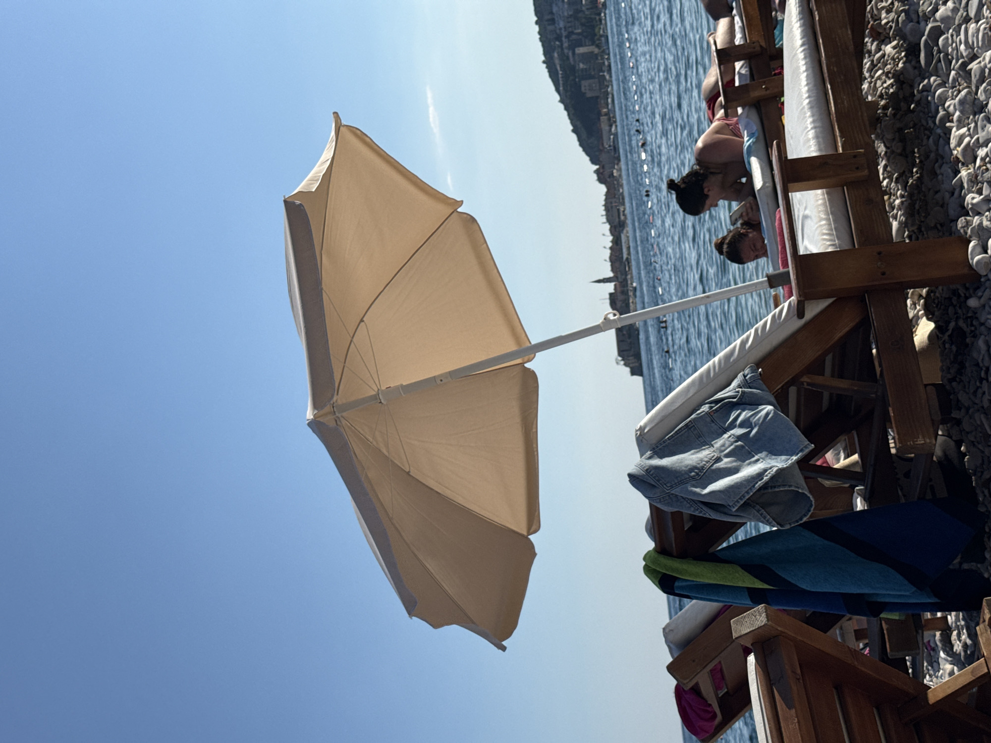 The image depicts a sunny day at a pebble beach in Budva, Montenegro. In the foreground, several wooden sun loungers are arranged on the pebbled shore, some with towels and clothing draped over them. A large, beige beach umbrella is set up, providing shade. In the middle ground, the calm, blue sea stretches towards the horizon, dotted with a few swimmers and a safety line. Across the water, the cityscape of Budva is visible, characterized by its historic buildings and a prominent church tower. Two women are relaxing on a sun lounger in the right side of the frame, one looking at her phone and the other facing away from the camera. The sky is clear and blue, indicating pleasant weather.