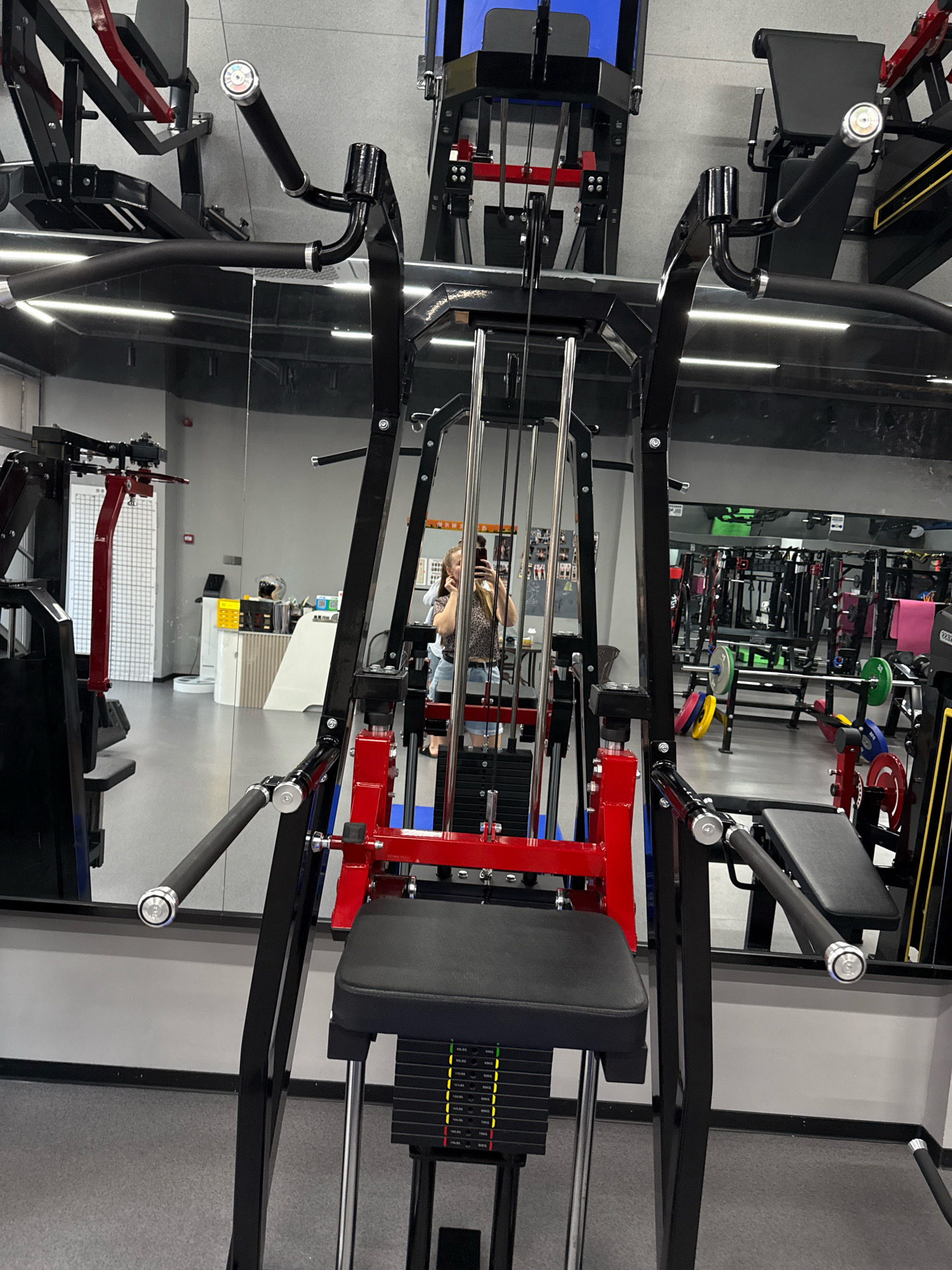 The image depicts an interior view of a modern gym, likely located in Yiwu, China, characterized by its clean, grey floors and wall finishes. The foreground is dominated by a black and red multi-station weight machine, featuring padded seats and weight stacks. To the left, another similar machine is partially visible. In the mid-ground, a person is captured in a reflection, holding a smartphone and appearing to take a selfie. The background showcases additional gym equipment, including weight benches, racks, and various weight plates in different colors, all arranged within the spacious gym environment. The overall lighting is bright, highlighting the polished surfaces of the equipment and the reflective qualities of the mirrors.