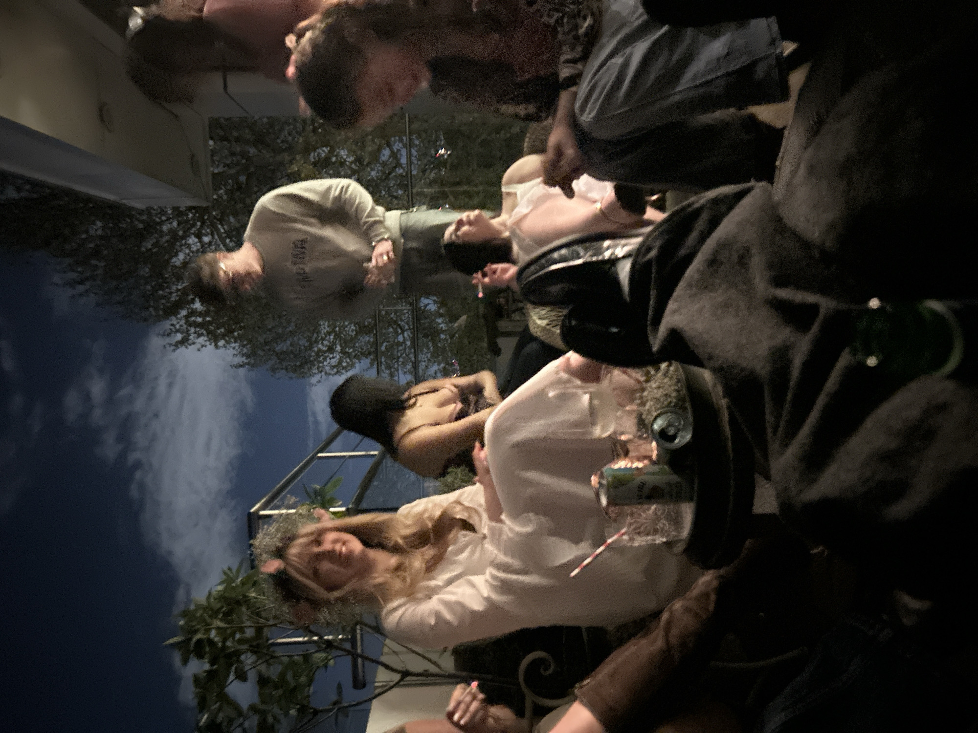 A group of people is gathered outdoors at night in what appears to be a social setting. The location is not discernible as being in London, United Kingdom based on the image alone. Several people are seated around a table with drinks visible. One woman in a white shirt has flowers in her hair. Another person is wearing a gray sweatshirt. Some individuals are partially obscured by the foreground elements. The lighting is dim and possibly from artificial sources. Vegetation is visible in the background.