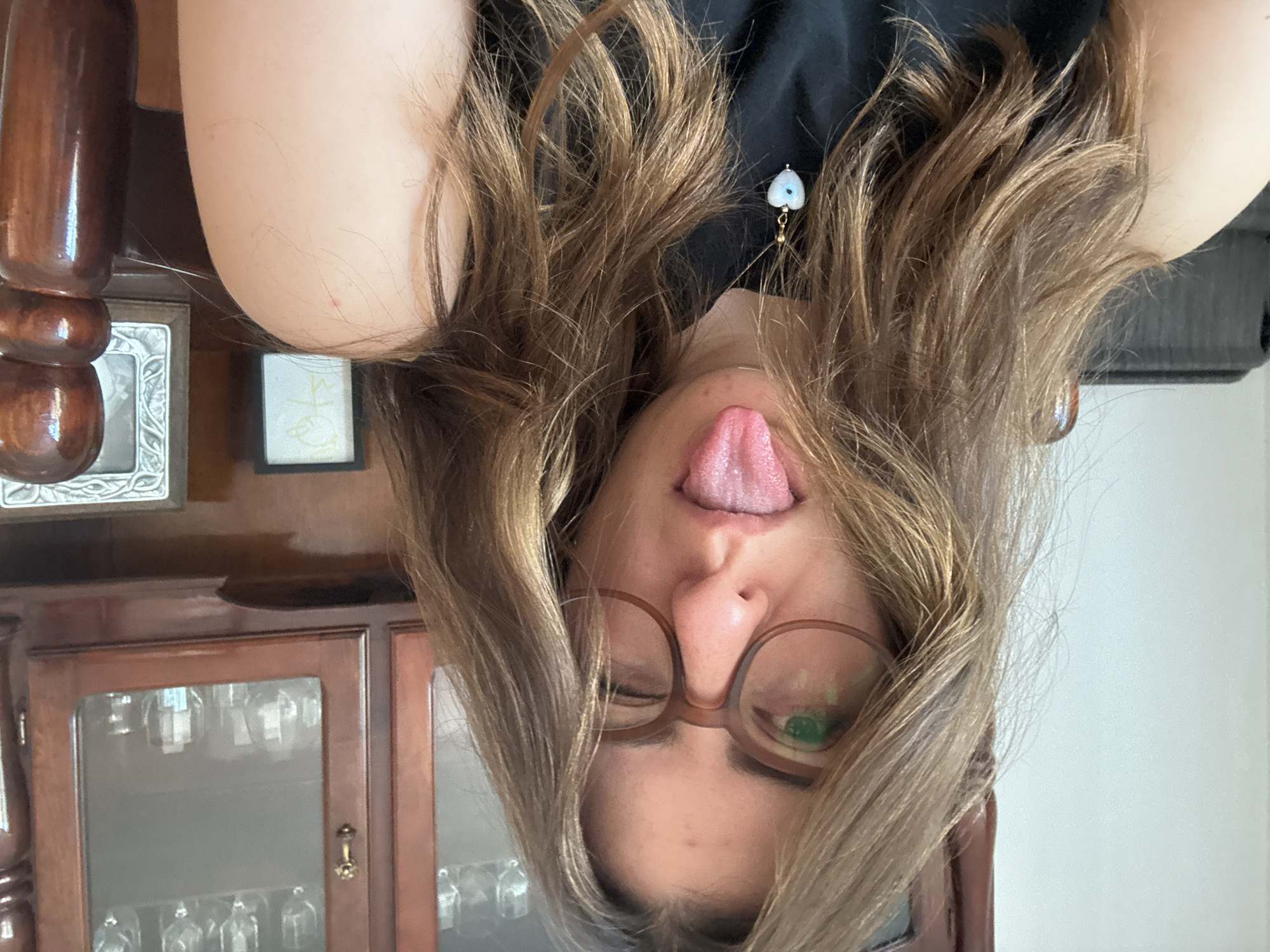 A person with brown hair, wearing glasses and a black top, is pictured upside down, sticking their tongue out. An earring with a light blue and white pendant is visible. The background includes a wooden cabinet, a framed picture, and a decorative wooden post. The location is Puebla, Mexico.
