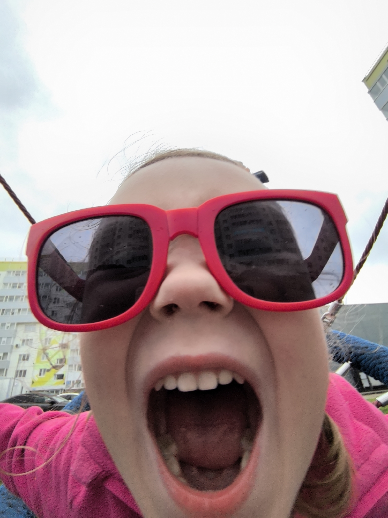 A close-up, low-angle, first-person perspective shot captures a young girl wearing oversized bright red sunglasses and a pink hooded jacket. Her mouth is wide open, revealing her teeth and the inside of her mouth, suggesting she might be shouting or laughing. Her blonde hair is visible, with a few strands falling forward. The background is out of focus and shows apartment buildings with windows, typical of a residential area in a city. The sky is overcast. The image appears to be taken on a swing set, with ropes visible on either side, and possibly in the city of Фролы, Russia, as suggested by the architecture.