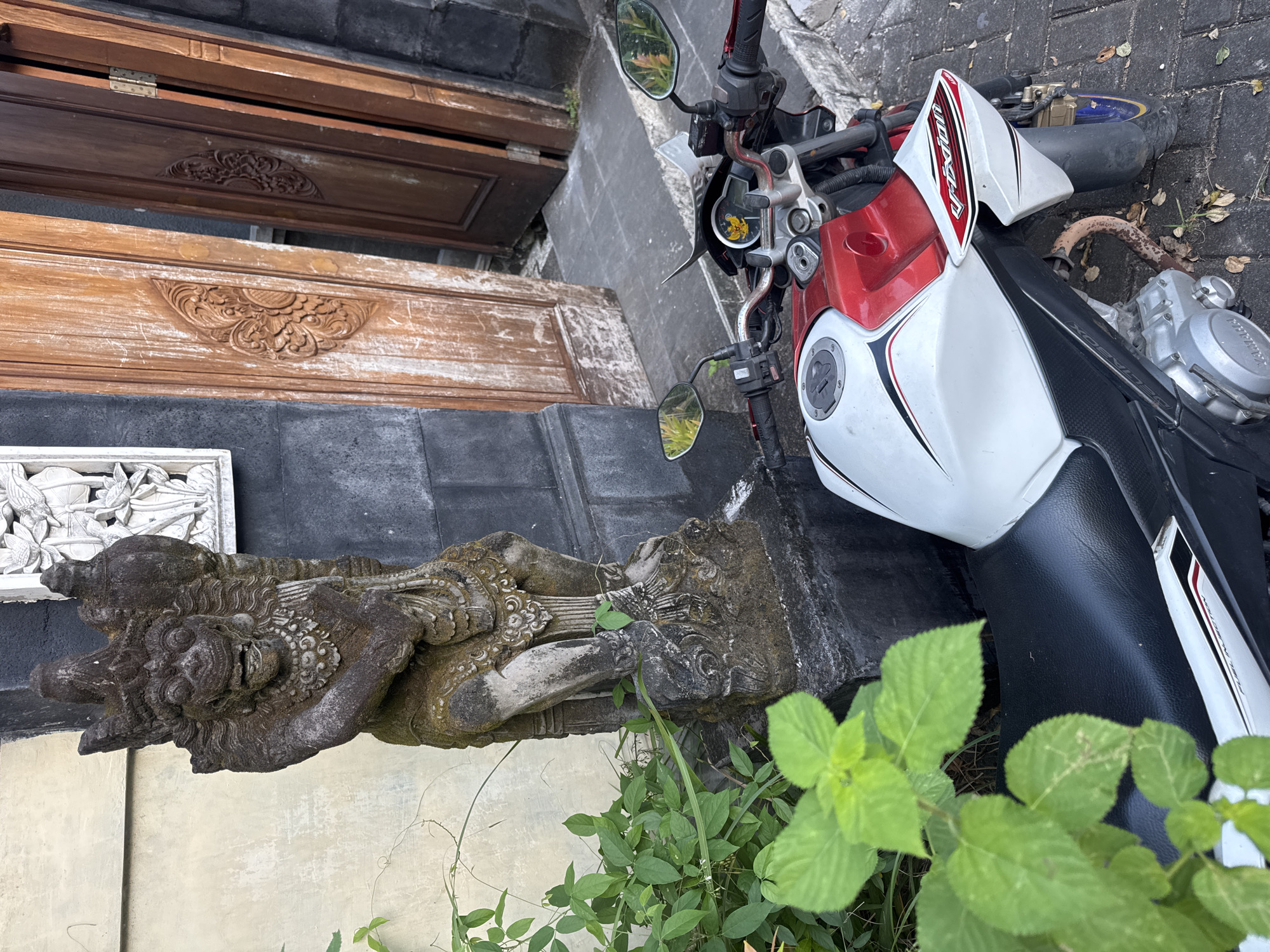 A motorcycle lies on its side next to a Balinese stone statue outside a building with carved wooden doors in Kuta Selatan, Indonesia. The motorcycle is predominantly white and red with a black seat. The statue appears weathered and covered with moss. Green foliage grows around the base of the statue. The building is built of dark brick or stone, with decorative panels above the statue and doorway. The surrounding ground is paved with stone.