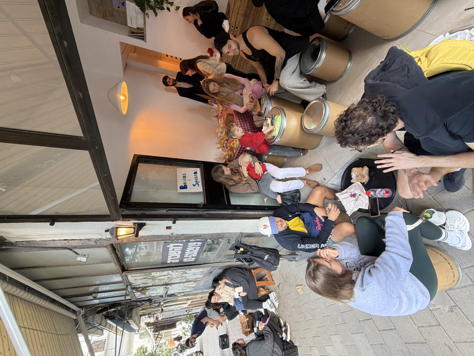 The scene depicts a lively gathering on a paved sidewalk and within the entrance of a storefront in Tel-Aviv, Israel. The building's exterior, visible on the left, features weathered plaster, large dark-framed windows, and a partially obscured sign in Hebrew reading "להשכרה" (For Rent) and the Tel-Aviv area phone number "03-5229219".
Approximately 15-20 individuals are present, varying in age from infants to adults. Seating is informal, consisting mainly of light brown industrial barrels with metal rims, repurposed as stools and low tables, and some wooden stools.
In the immediate foreground, a woman wearing a grey sweatshirt and dark green pants sits on a barrel, facing a man with dark curly hair wearing a black top and yellow garment, who is seated on the ground and gesticulating with both hands. A small black table between them holds a water bottle, a mobile phone, and some discarded paper.
To their left, another man in a white baseball cap, dark jacket, and a blue apron-like garment sits on a barrel, holding a cup. A black bicycle is parked nearby. Further left, along the sidewalk, several people are seated on stools or barrels, including a woman holding a large brown wicker bag.
Inside the storefront entrance, marked by a sign stating "BREADS & SPREADS," a red and white checkered tablecloth covers a display table laden with various baked goods and small bowls of spreads. A woman in a brown top and white pants sits on a barrel, holding a fair-haired child dressed in red. Next to them, another child in a red top interacts with items on a barrel used as a table. Other individuals are seated or standing inside, including a man with a beard wearing a dark suit in the background and a woman sitting on a raised platform to the far right. The interior is brightly lit by a pendant lamp and a wall sconce illuminates the entrance.