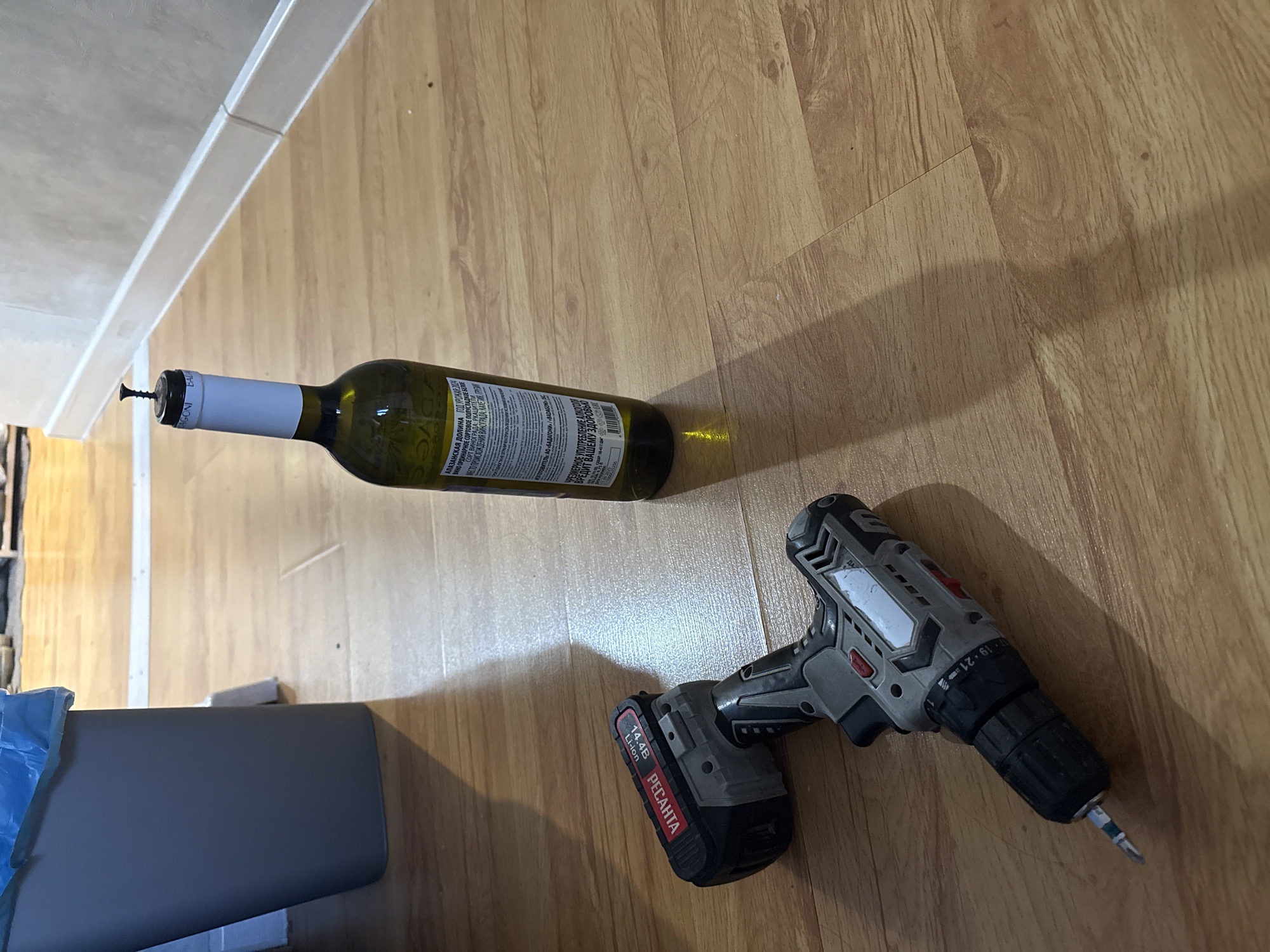 The scene shows a partially consumed bottle of "Alazani Valley" white wine lying horizontally on a light brown, wood-patterned floor, next to a gray and black "Ресанта" (Resanta) cordless drill. A screw is stuck in the neck of the bottle. The wine label indicates the wine is bottled in Balagoni (Балагони), Russia. The OCR text identifies the wine as ordinary white semi-sweet, made from Rkatsiteli grapes. There is a corner of a light gray object with blue wrap partially visible, possibly a piece of furniture, in the lower left corner of the image and a wall running along the top left side. The Izhevsk, Russia location cannot be confirmed.