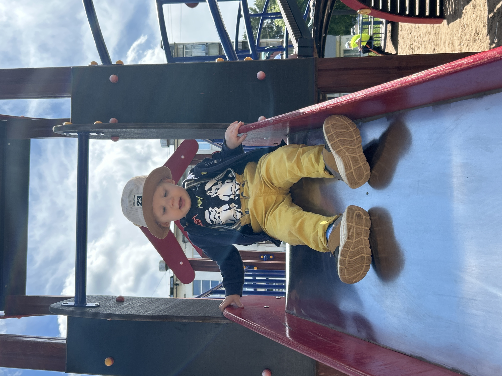 A young child wearing a bucket hat, dark jacket with Star Wars characters, and yellow pants is sitting on a metal slide at a playground. The child is looking down the slide. The playground structure appears to be made of wood and metal. In the background, there are buildings, trees, and other playground equipment. The sky is cloudy. The location is in Chaikovsky, Russia.