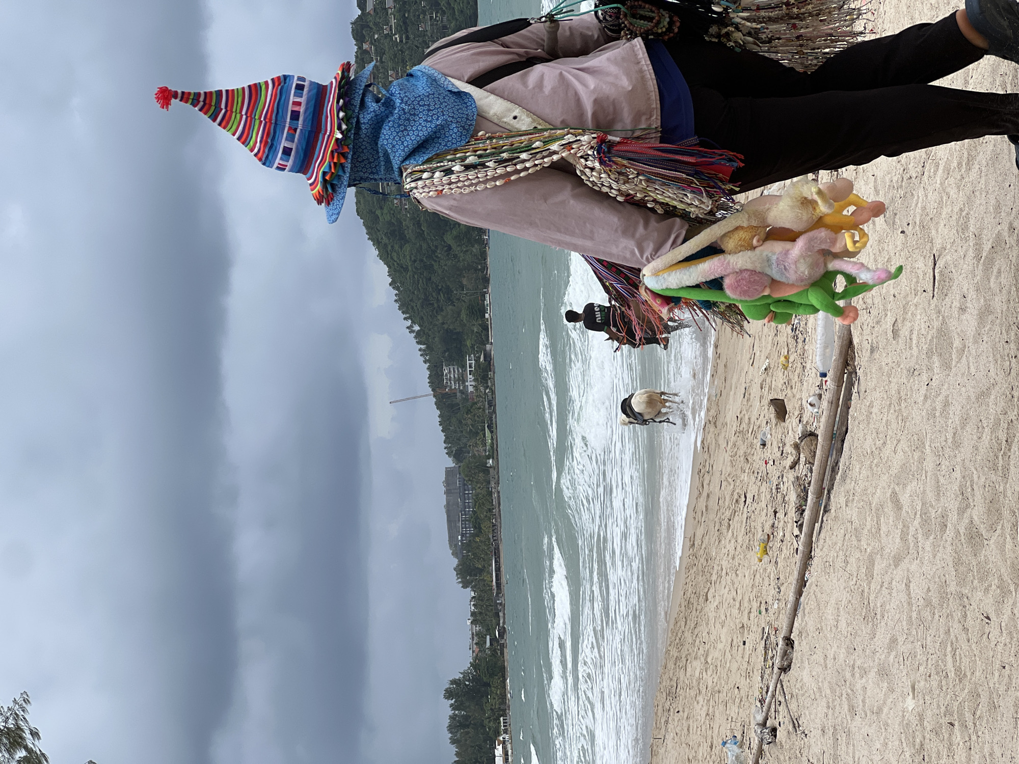 A vendor, seen from behind, walks along a sandy beach in Choeng Thale, Thailand, carrying several plush toys and necklaces. The vendor wears a colorful, conical hat, a blue face covering, and a light-colored jacket over dark pants. In the background, the ocean has large waves, and a person is riding a horse in the surf. Buildings and lush green hills are visible in the distance, under a cloudy sky. The foreground of the beach is littered with various debris, including plastic bottles and driftwood.