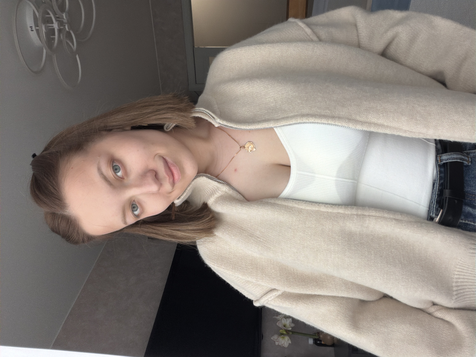 A close-up, slightly tilted portrait captures a young woman, likely in her late teens or early twenties, looking directly at the camera with a subtle, pleasant smile. She has light brown hair, styled straight, and striking blue-green eyes. She is dressed in a casual yet chic ensemble, featuring a light beige or off-white knitted cardigan or jacket, partially zipped, revealing a white ribbed tank top or camisole underneath. A delicate gold-toned necklace with a circular pendant, possibly depicting a stylized flower or geometric design, adorns her neck. The lower part of the image shows a glimpse of dark blue jeans and a black belt on her right side.
The setting is indoors, appearing to be a contemporary room in Reshetnikovo, Russia. The background consists primarily of a light gray wall. In the upper left, a modern, artistic ceiling light fixture with multiple transparent or white circular elements is visible. Towards the lower right, a dark surface, possibly a table or shelf, is partially seen, decorated with a small arrangement of white flowers in a vase. The lighting is bright and even, indicating an indoor shot, possibly a selfie or a close-up portrait. No text is visible in the image.