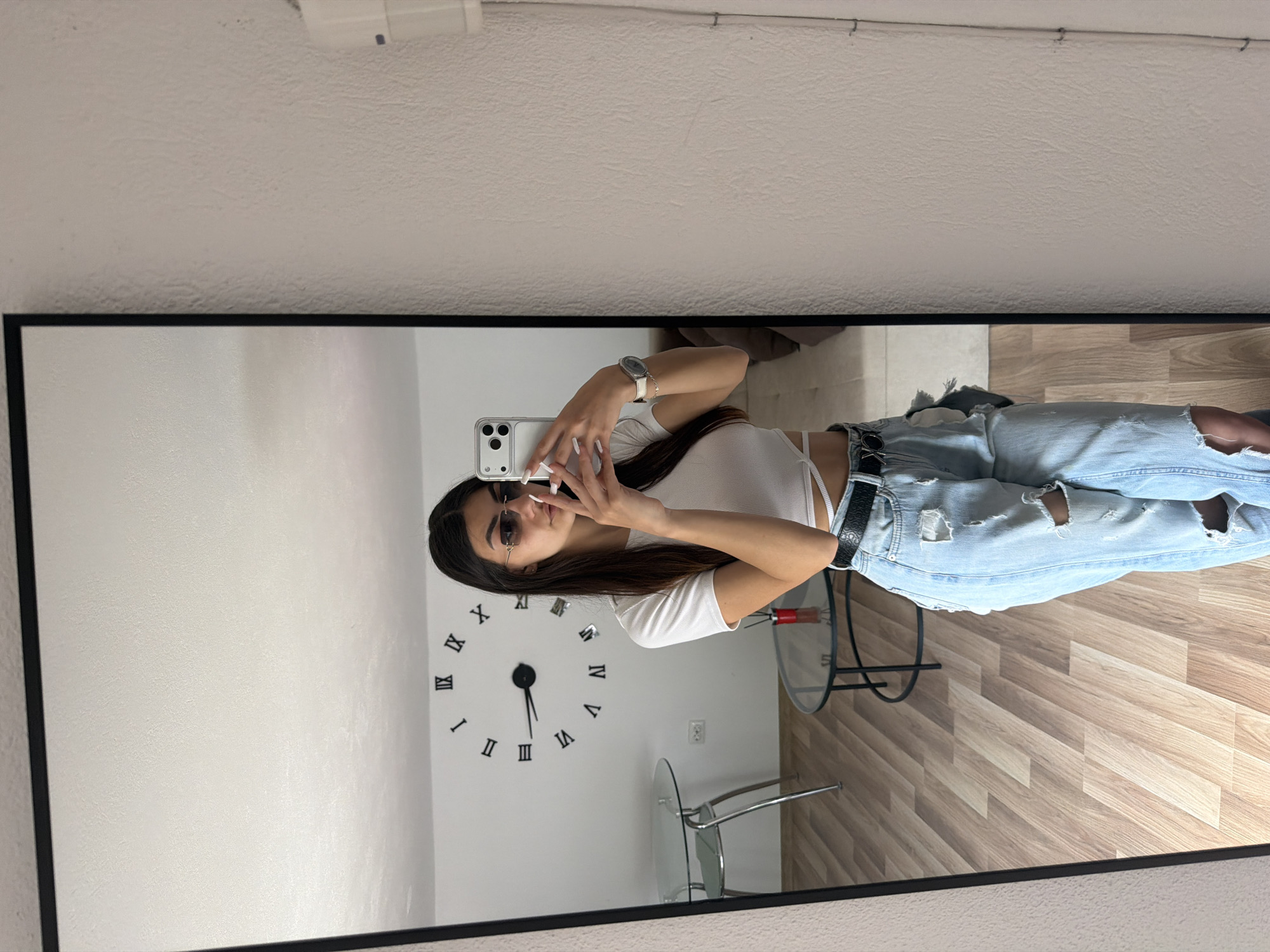 A woman is taking a selfie in a mirror. She has long brown hair and is wearing a white top, ripped blue jeans, sunglasses, and a watch. She is holding a white smartphone. Behind her in the reflection is a white wall with a decorative clock on it, two small glass tables with metal frames, and a wood-patterned floor. The photo appears to be taken indoors. The city of Gabrovo, Bulgaria cannot be determined from the image.