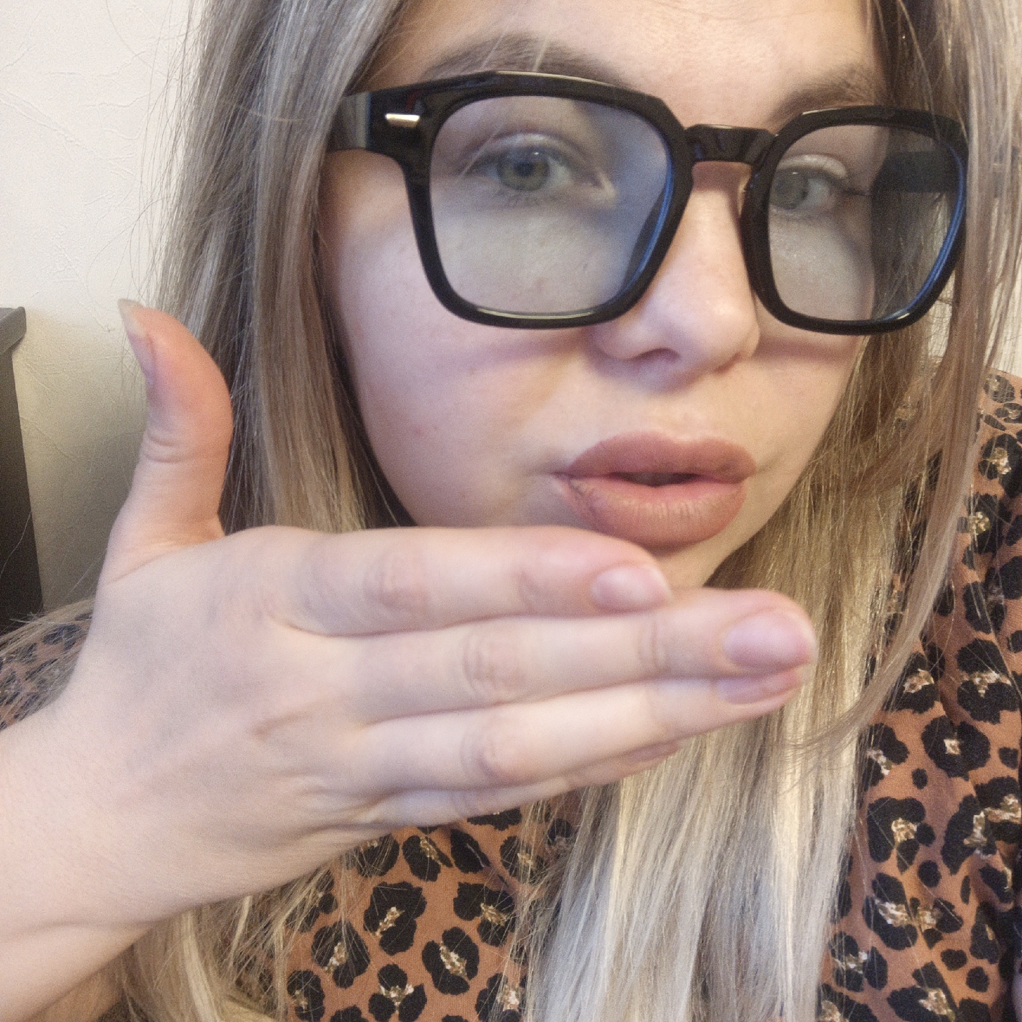 A close-up, eye-level shot features a woman looking directly at the camera, posing as if blowing a kiss. She is wearing large, black, square-framed glasses that obscure parts of her eyes with a slight reflection. Her eyes, visible through the lenses, appear to be green or hazel. Her lips are slightly puckered, suggesting a kiss, and are adorned with a natural, light-toned lipstick.
Her long, ash blonde or light brown hair frames her face and flows down her shoulders. She is wearing a top with a distinctive brown and black leopard print pattern, visible on her right shoulder and chest area. Her left hand is raised in front of her face, palm facing the camera, with fingers slightly curled, as if delivering or catching a kiss. Her nails are short and natural. The background on the left consists of a plain, light-colored wall, indicating an indoor setting. The lighting is soft and even, suggesting either diffused natural light or indoor artificial lighting. No text is visible in the image.