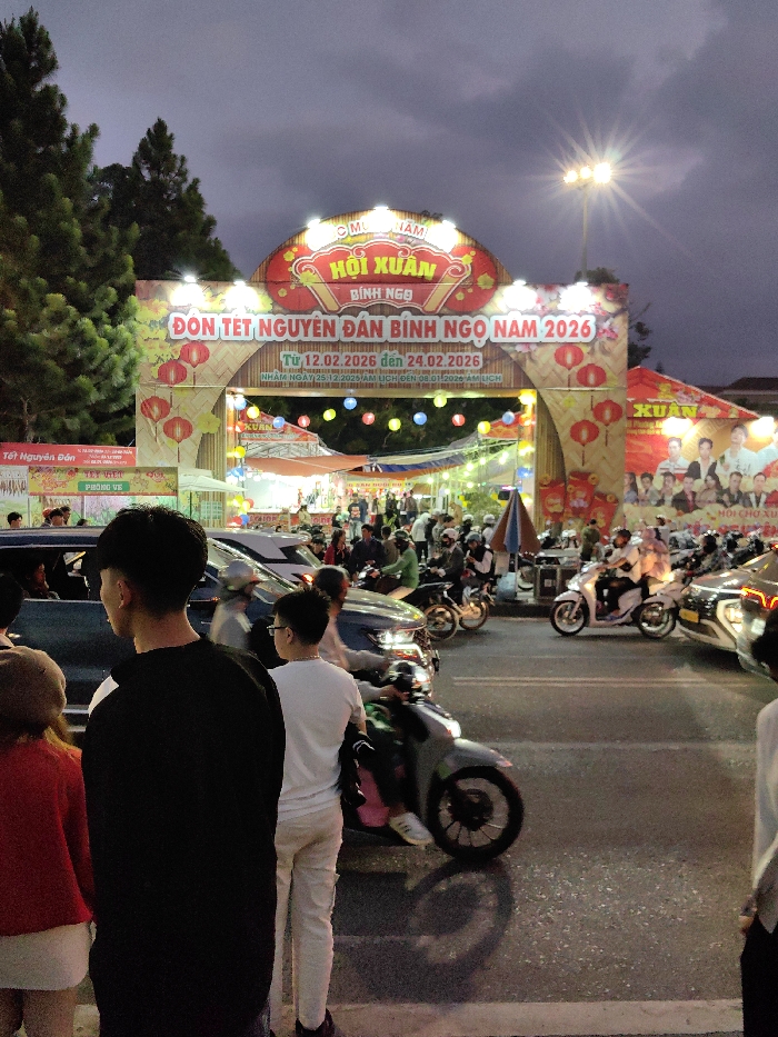 The image captures an evening scene at the entrance of a Tet (Lunar New Year) festival in Da Lat, Vietnam. A large, brightly lit archway dominates the central view, featuring Vietnamese text that reads "HỘI XUÂN" (Spring Festival) and "ĐÓN TẾT NGUYÊN ĐÁN BÍNH NGỌ NAM 2026" (Welcome Tet Nguyen Dan Binh Ngo Year 2026), with event dates specified from February 12, 2026, to February 24, 2026. The archway is decorated with numerous white lights, red lanterns, and a bamboo-style motif.
Behind the archway, a bustling outdoor market or festival area is visible, illuminated by string lights with various colorful bulbs and overhead awnings or tents. Dark green pine trees are present on the left side of the frame, behind the archway. The sky above is dark and cloudy. A tall streetlight on the right side emits a bright starburst effect.
In the immediate foreground, three individuals are partially visible from the back or side. From left to right, these include a person in a red top and light skirt, a male in a black long-sleeved shirt with white sleeve accents, and a male in a white t-shirt and glasses.
The paved roadway in the midground is densely populated with a mix of vehicles and pedestrians. Numerous motorcycles and scooters, many with riders wearing helmets, are present, some appearing to be in motion or queued. Several cars are also visible, some with active headlights. The scene depicts a lively public gathering during a festival.