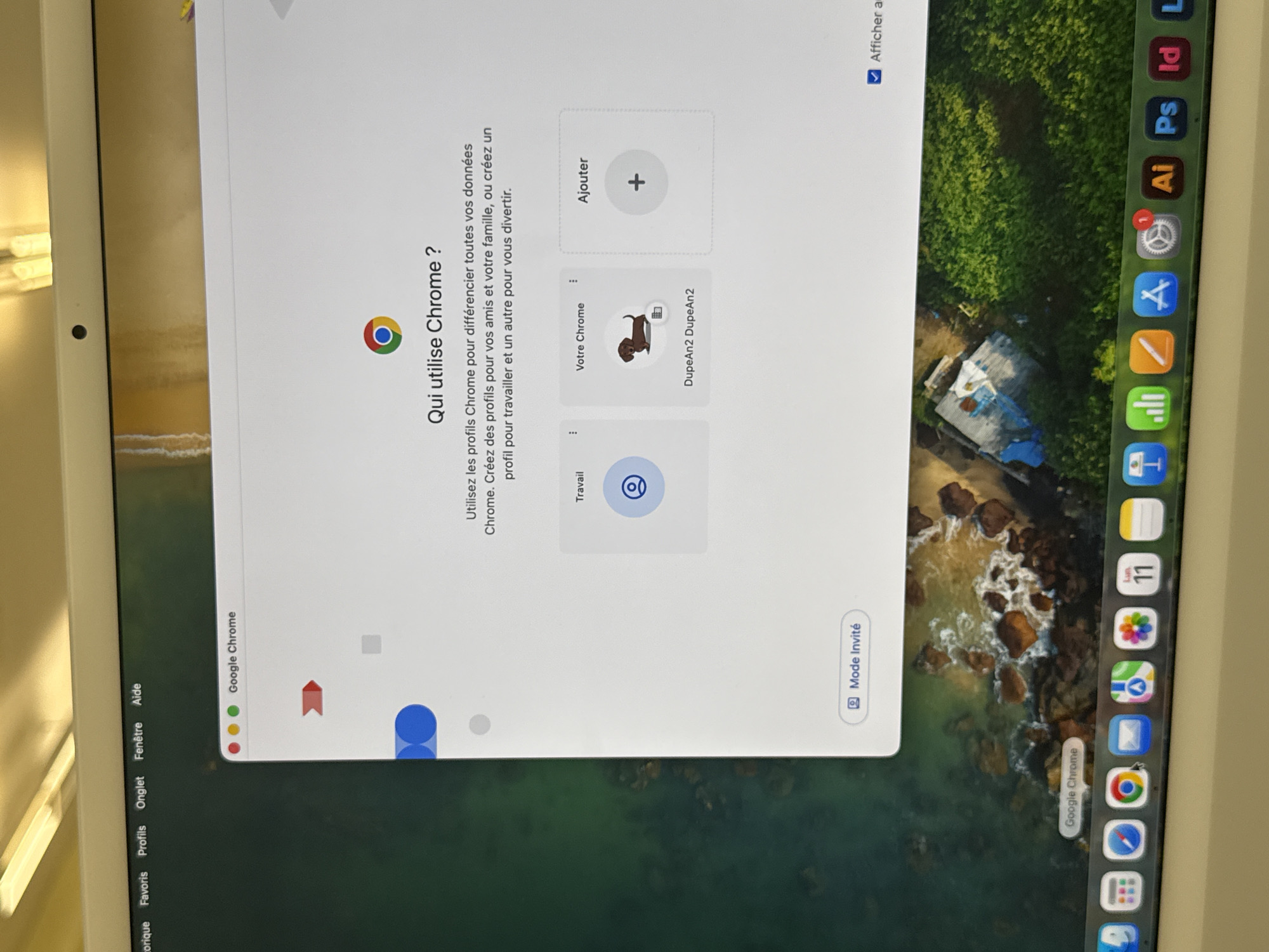 The image shows a laptop screen displaying the Chrome browser window. The Chrome window asks "Qui utilise Chrome?" (Who uses Chrome?) and explains that Chrome profiles can be used to differentiate data for friends, family, or work. There are options to select a "Travail" (Work) profile, "Votre Chrome" (Your Chrome) profile named "DupeAn2 DupeAn2", or to "Ajouter" (Add) a profile.
The laptop's dock is visible at the bottom, showing icons for various applications, including Google Chrome, Safari, and Adobe applications like Illustrator (Ai), Photoshop (Ps), and InDesign (Id).
The desktop background is a satellite image of a tropical area including a small town and the ocean. The image is taken indoors. The presence of French language indicates that the locale is most likely Quebec, Canada. It cannot be confirmed that the location is Montreal.