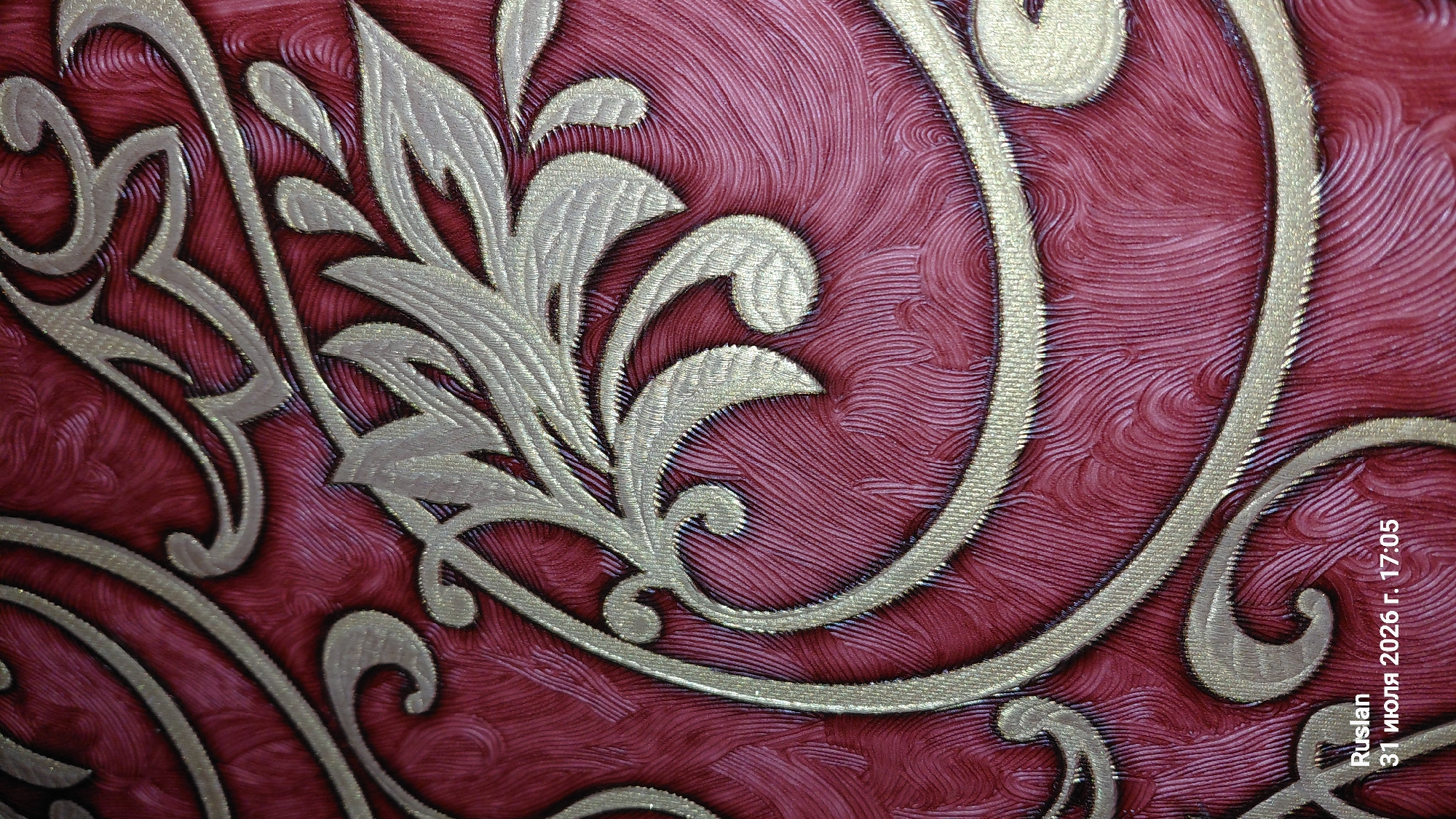 The image is a close-up, low-angle shot of ornate red and gold wallpaper. The wallpaper features a swirling, embossed pattern of floral motifs and scrollwork in a metallic gold finish against a textured crimson background. The texture of the red background appears to be a series of fine, wavy lines. The bottom left corner of the image displays text that reads "Ruslan" followed by a date and time: "31 июля 2026 г. 17:05". The location of Oleksandriia, Ukraine is not depicted in the image itself; the text appears to be a watermark or overlay.