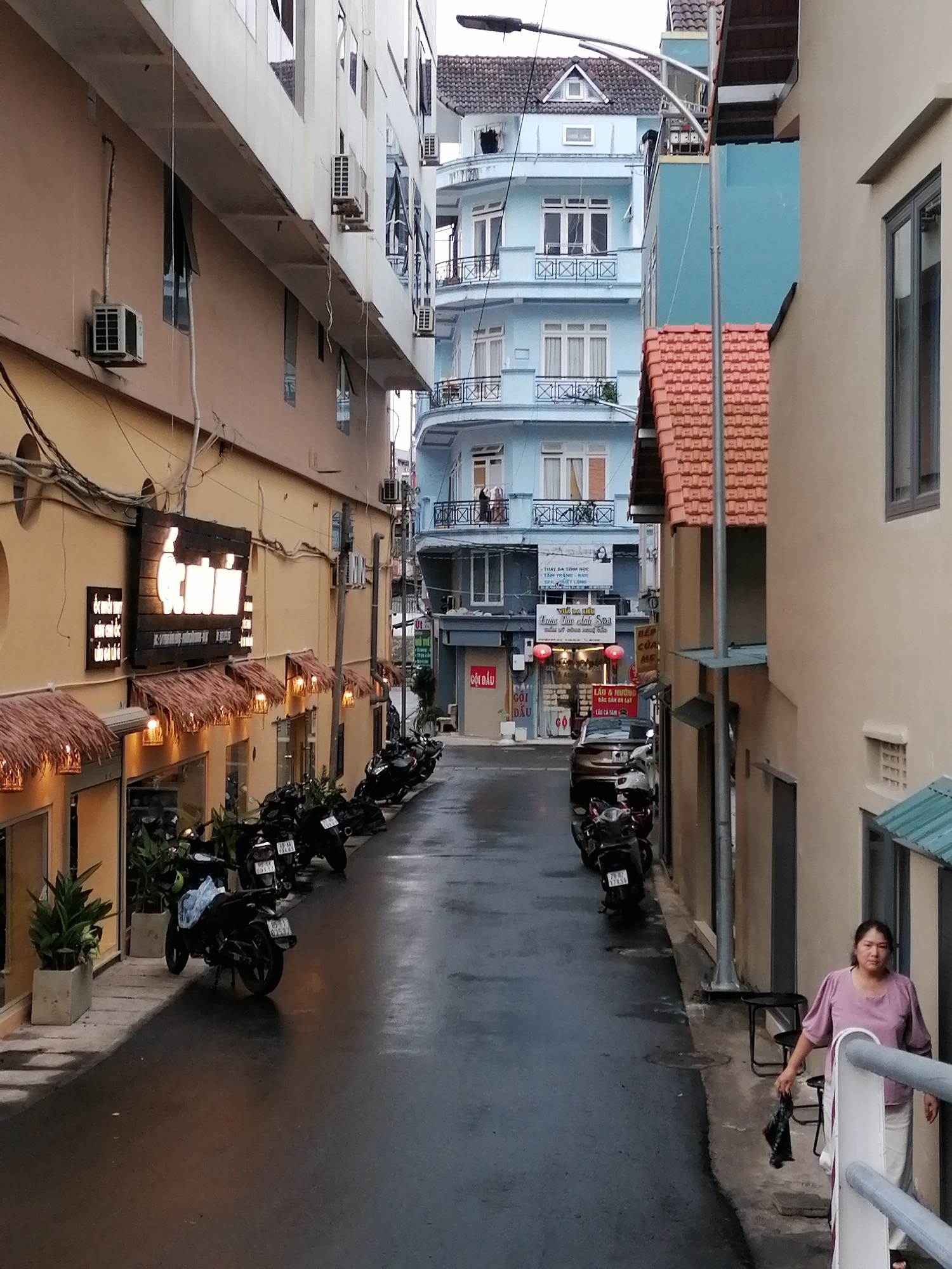 This is a medium-angle, eye-level shot of a narrow street in Da Lat, Vietnam, likely taken in the late afternoon or early evening. The wet asphalt street suggests recent rain, and the dim lighting contributes to a somewhat subdued atmosphere. The scene is flanked by multi-story buildings, typical of Vietnamese urban architecture, with a mix of modern and older styles.
On the left side of the street, a row of shops with thatched awnings and illuminated signs are visible. The signs display Vietnamese text, including "GỘI ĐẦU" (hair washing) and "SPA," indicating commercial establishments. Several motorcycles are parked along the sidewalk, a common sight in Vietnam. Potted plants add touches of greenery to the otherwise concrete surroundings.
On the right, a woman in a light purple shirt and white pants stands near a railing, holding a dark bag. Her expression is neutral. The building on this side is lighter in color and features a red-tiled roof. Further down the street, more buildings are visible, including a striking light blue structure with multiple balconies. The overall scene captures a slice of everyday life in a bustling Vietnamese city, with a focus on the street's commercial activity and the presence of parked motorcycles.