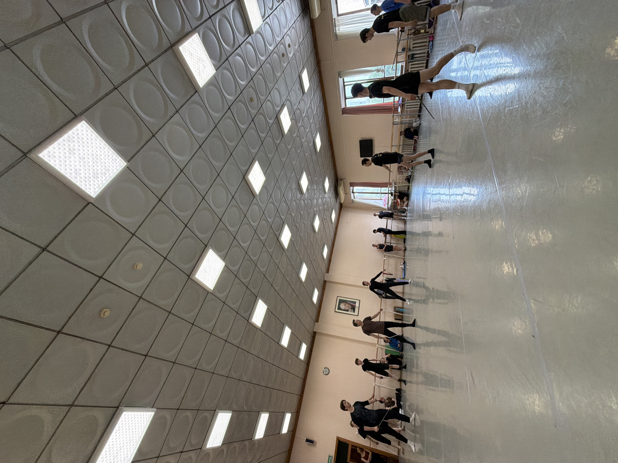 The image captures an indoor ballet class in progress, possibly at a dance studio within Ufa, Russia. The room is spacious with a polished floor reflecting the light. A prominent feature is the acoustical tile ceiling with embedded rectangular fluorescent lights and circular textured patterns. Ballet barres run along the wall, indicating a dedicated dance space. Several young men are actively participating in the class, dressed in athletic wear, and some are holding thin rods, likely for practice. Others are seated or observing, suggesting a learning environment. The walls are painted a light, neutral color, and a framed portrait is visible on one wall, alongside a door marked with an exit sign in Russian. Windows with light-colored curtains allow natural light to enter the room, contributing to the bright and airy atmosphere. The overall scene depicts a disciplined and focused training session.
