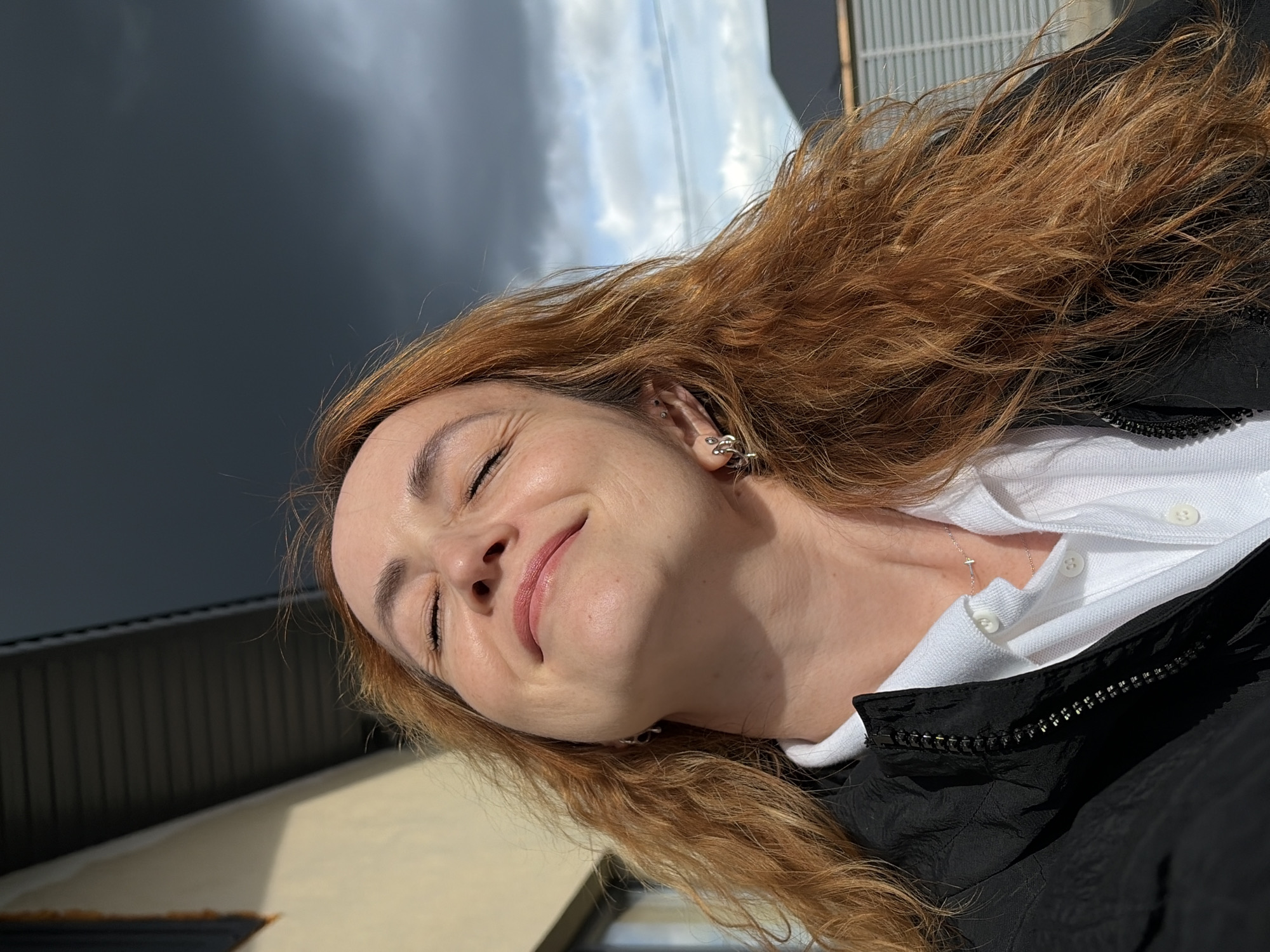 The image shows a close-up portrait of a woman with long, reddish-brown, wavy hair, tilted diagonally within the frame. Her eyes are closed, and a gentle, serene smile plays on her lips, suggesting contentment or enjoyment of the sunlight. She is wearing a white polo shirt beneath a black zippered jacket. A delicate silver necklace with a small, minimalist pendant is visible around her neck, and she has an intricate silver ear cuff or earring in her visible left ear.
The scene is illuminated by strong natural light, likely direct sunlight, casting bright highlights on her hair and face and creating defined shadows. The background consists of a partially cloudy sky, with areas of bright blue mixed with darker gray clouds. Below the sky, faint details of what appear to be building structures or fences with horizontal elements are visible. The setting is outdoors during the daytime, in Chaikovsky, Russia, under sunny and partly cloudy conditions. There is no visible text in the media file.