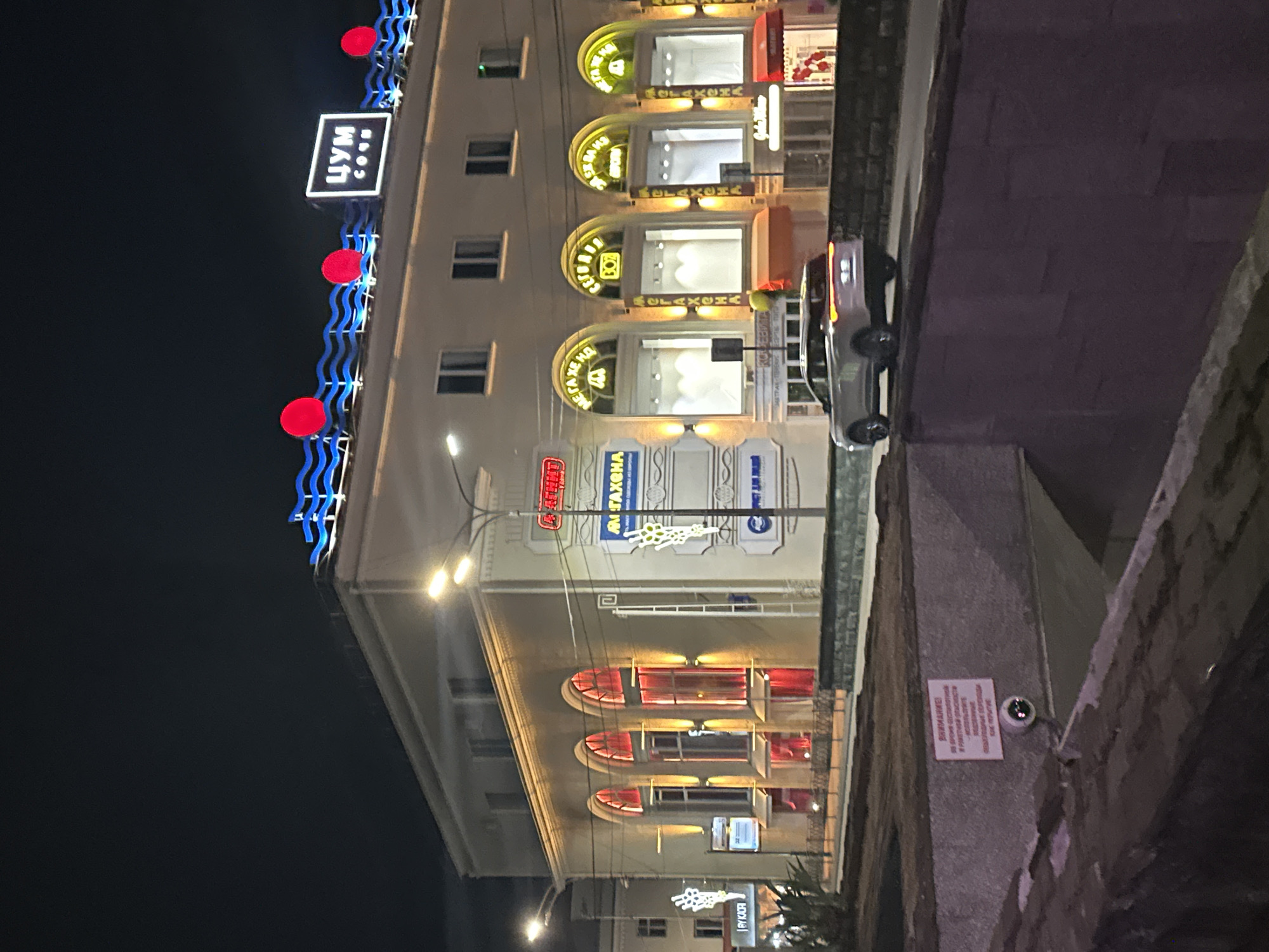 This image depicts the front facade of the TsUM department store in Sochi, Russia, at night. The building is illuminated by a combination of interior lighting, exterior spotlights, and prominent neon signage. The most striking features are the wavy blue neon lights along the rooftop, punctuated by red circular lights. Above the main entrance, a white rectangular sign with blue Cyrillic lettering reads "ЦУМ СОЧИ" (TSUM SOCHI).
The ground floor of the building features large arched windows, many of which are lit from within, with red curtains visible behind some of them. The windows are framed by decorative elements and signage, including yellow neon lettering that reads "МЕГАХЕНА" (MEGAKHENA) in several instances, likely indicating shops or brands within the department store. Above the ground floor are several smaller, rectangular windows on the second floor.
In the foreground, a light-colored SUV is driving on a road in front of the building. To the left of the building, a pedestrian walkway with a metal railing is visible. On a concrete wall in the lower left portion of the image, a surveillance camera is mounted next to a red and white sign with Cyrillic text. The overall environment is urban and suggests a commercial district. The lighting conditions indicate it is nighttime, with artificial lights illuminating the scene. No people are clearly visible.