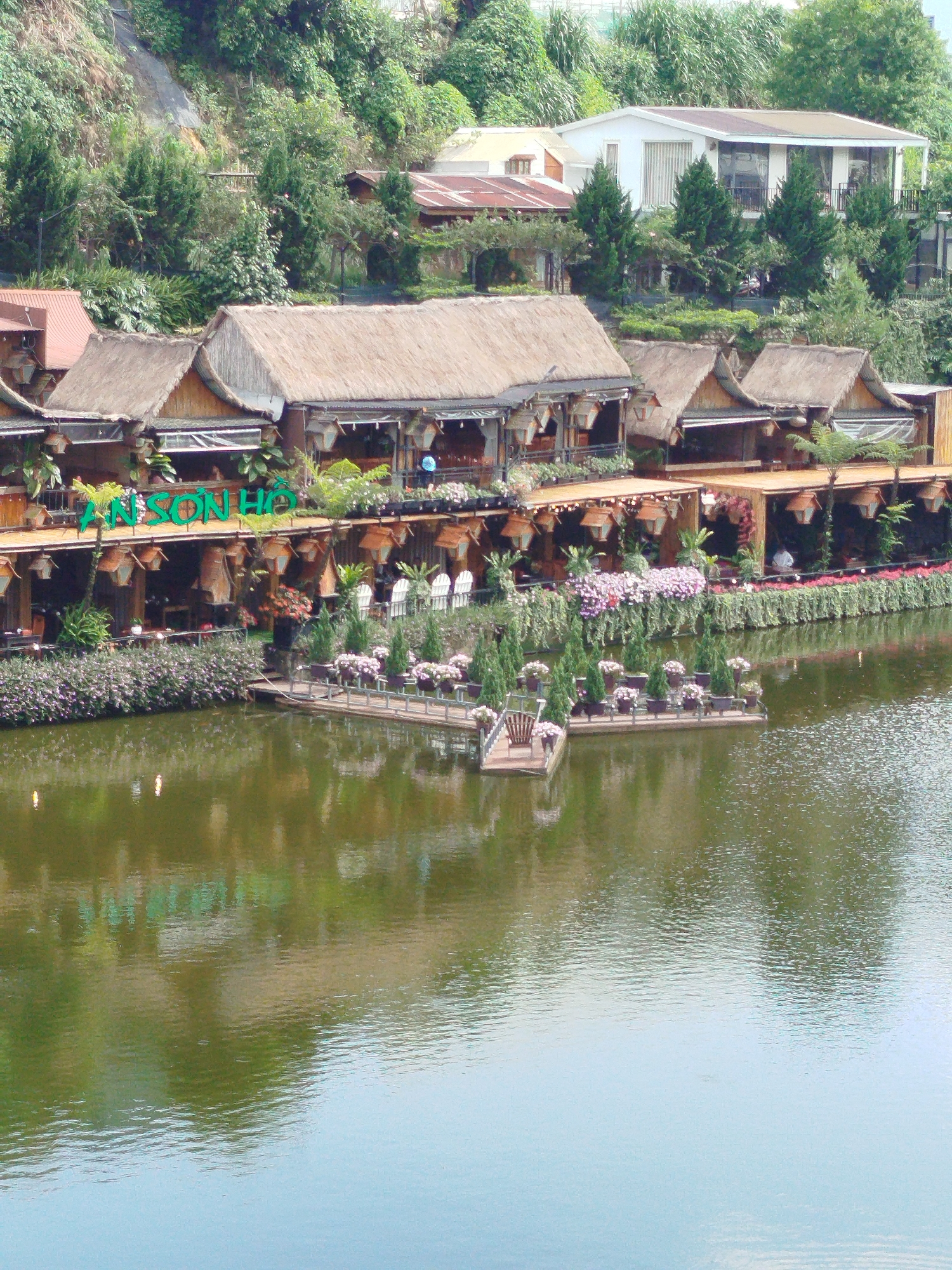 The image captures a scenic view of An Son Lake in Da Lat, Vietnam. A series of connected buildings with thatched roofs line the waterfront. The name "AN SON HO" is displayed in green lettering on one of the buildings. Below the buildings, a line of vibrant purple and red flowers adds color.
In the foreground, a small, floating platform with potted plants and a wooden chair extends into the lake. The water's surface reflects the surrounding architecture and foliage.
Behind the buildings, lush greenery and vegetation cover a hillside. A house with a mix of modern and traditional architectural elements sits atop the hill, partially obscured by trees.