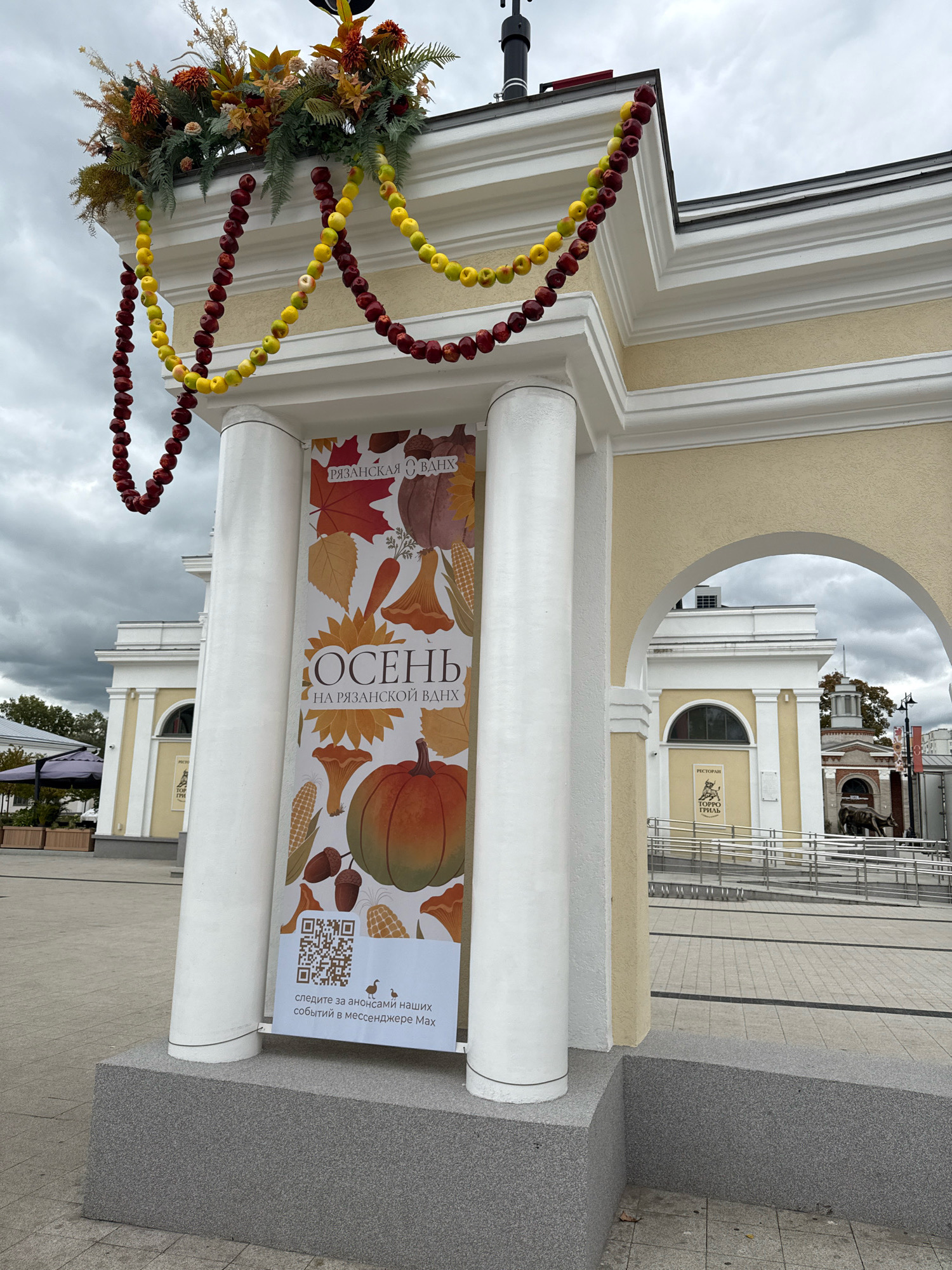 This image shows an outdoor scene in Ryazan, Russia, with a focus on a banner and autumnal decorations. The banner, featuring autumn-themed illustrations like pumpkins, leaves, corn, and mushrooms, is hung between two white pillars. The text on the banner reads "ОСЕНЬ НА РЯЗАНСКОЙ ВДНХ" which translates to "Autumn at Ryazan VDNKh". Below the illustrations, there is a QR code and text in Russian, which appears to be an invitation to follow announcements of events in a messenger.
Above the banner, there are garlands made of apples, in red and yellow varieties, and a floral arrangement. The architecture visible in the background consists of light-colored buildings with arched entrances and classical elements, characteristic of exhibition or cultural centers. The overall atmosphere suggests an event or celebration related to autumn. The sky is overcast, indicating a cloudy day. There are no people clearly visible in the immediate foreground.