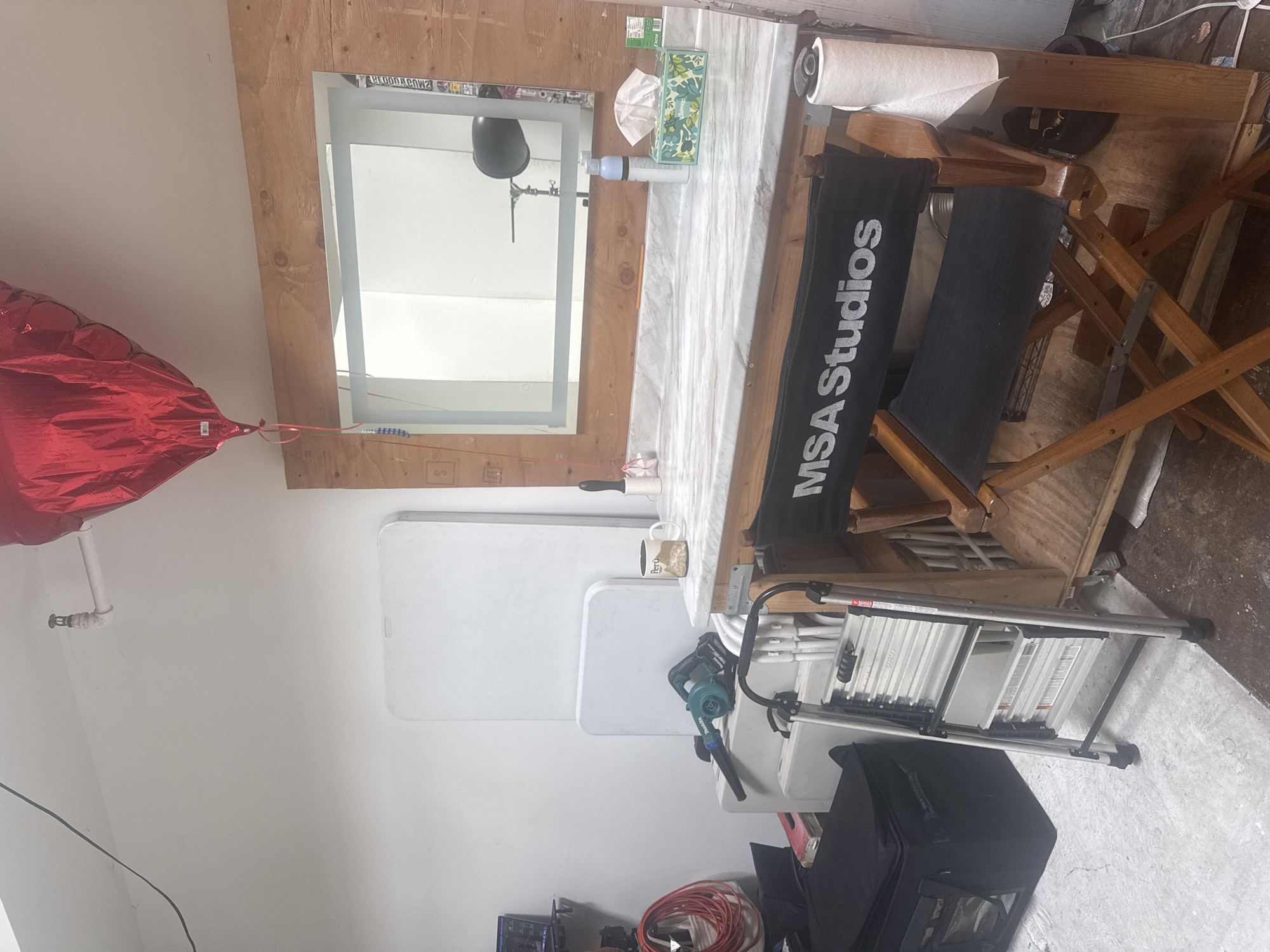 The image depicts an indoor scene, possibly a workshop or studio space. A red balloon is visible on the left side of the image, tied to a wall. A director's chair with the words "MSA Studios" printed on it is situated next to a table. On the table, there are various items, including a box of tissues and a roll of paper. A mirror is mounted on the wall behind the table. Beneath the table, there are white plastic folding tables stacked. A leaf blower and a metal stepladder are present near the tables. A black bag is located on the floor. Based on the "MSA Studios" on the chair, the location could potentially be in Los Angeles, United States of America, as MSA Studios has locations in that city.