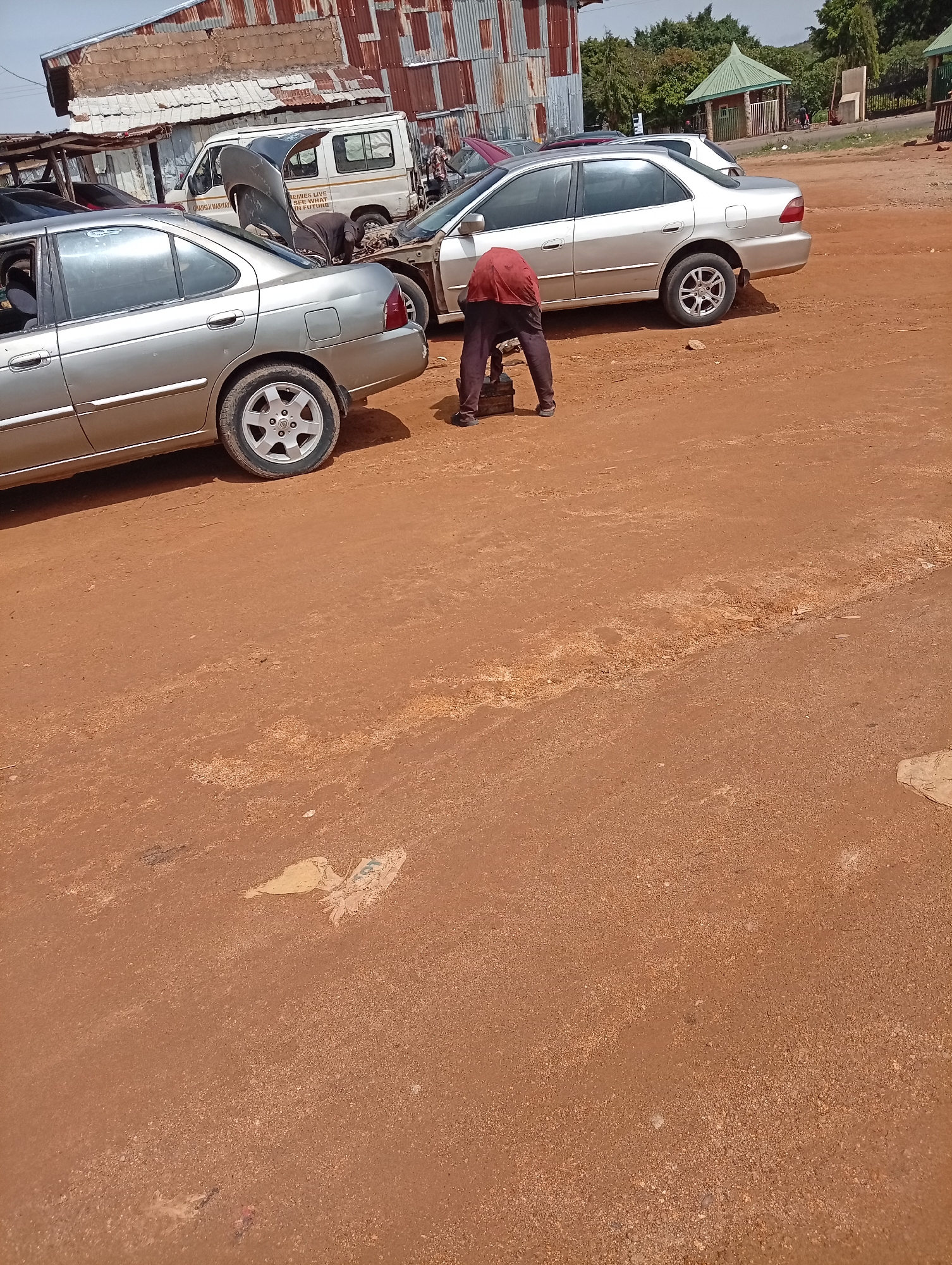 The image depicts an outdoor scene in Jos, Nigeria, likely a roadside auto repair area. A man in a red shirt and dark pants is bending over near the front of a silver sedan. The sedan has its hood open, suggesting engine work. To the left, another silver sedan is parked with the trunk slightly ajar. A white van with writing on its side is behind the cars, and another maroon car is partially visible. The ground is reddish-brown dirt, with a light-colored patch in the lower-center of the image. In the background is a building with a corrugated metal roof and a green-roofed structure to the right.