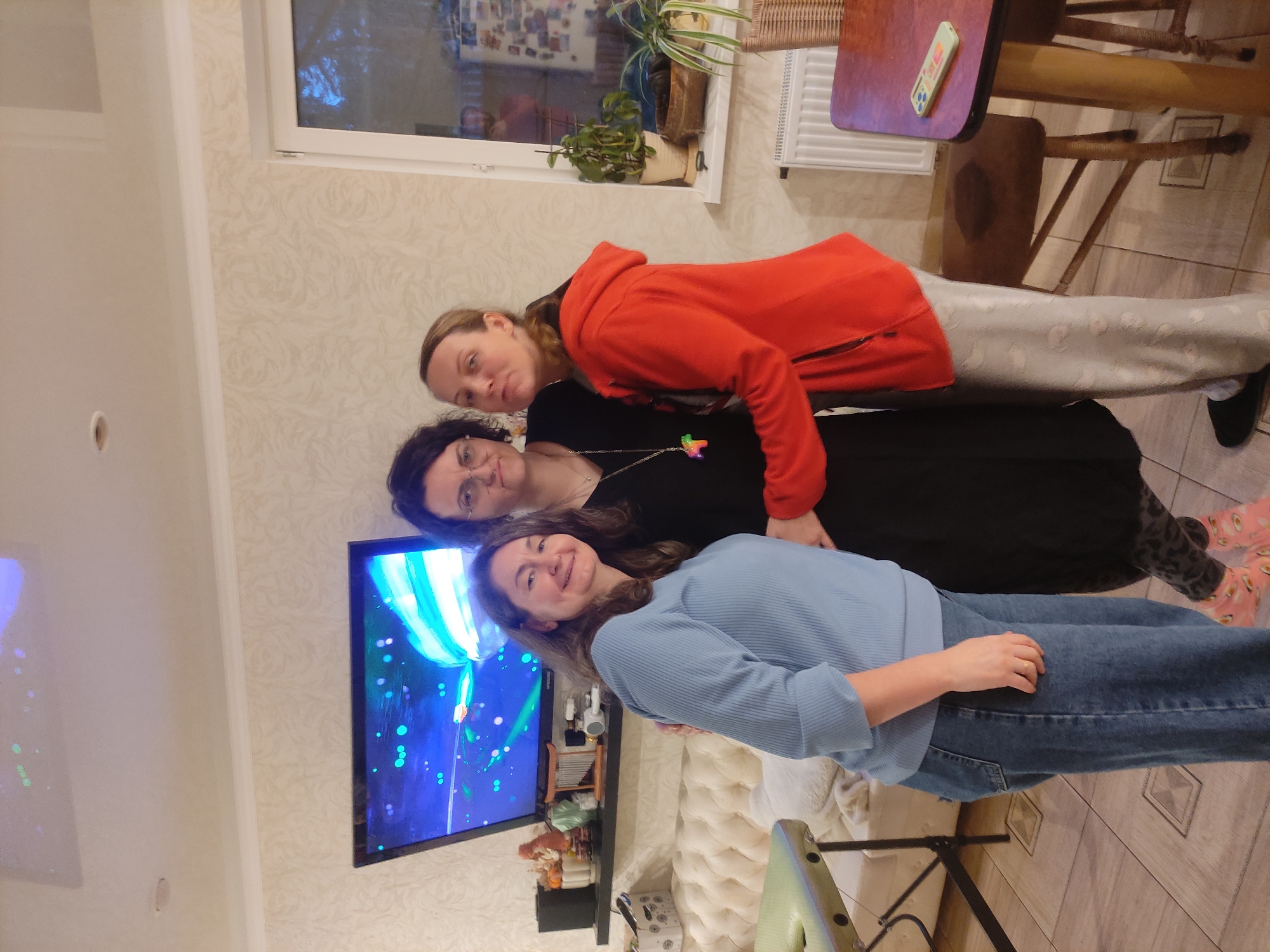 Three women are posing indoors. The woman at the top is wearing a red hoodie and light gray pants. The woman in the middle has dark curly hair and glasses and is wearing a black dress. The woman at the bottom has medium brown hair and is wearing a light blue sweater and jeans. They are posed in front of a wall with patterned wallpaper. A television is visible on the wall to their left, displaying an image with blue, green, and white colors. To the right, there is a wooden table with a remote control on it. There is also a chair and a radiator near the table. Plants in pots are sitting on the window sill. The floor is tiled. The location is an interior space within a house. The city is Unknown, Unknown.
