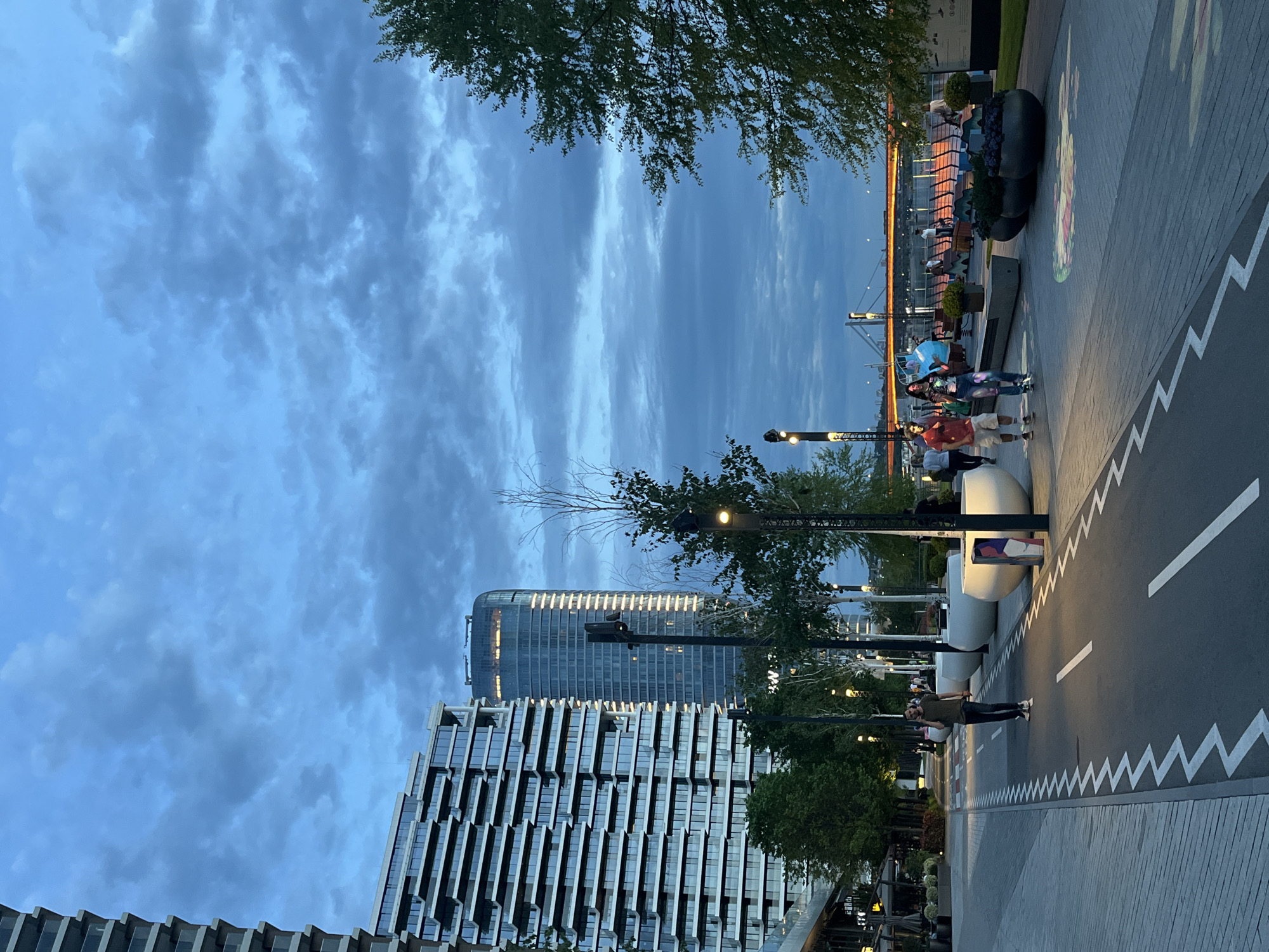 The image shows a modern urban scene in Belgrade, Serbia. The perspective is along a paved walkway with stylized white zigzag and linear markings, resembling a road design. There are two large, contemporary buildings on the left, one with a multi-story, horizontal glass façade, and another taller tower behind it. Several young trees line the walkway, some with lit fixtures. People are visible along the path: some standing, others walking. Beyond the walkway is an elevated platform with a metal railing, where more people are visible. Large planter boxes are placed along the platform. The sky is overcast with blue and grey clouds.
