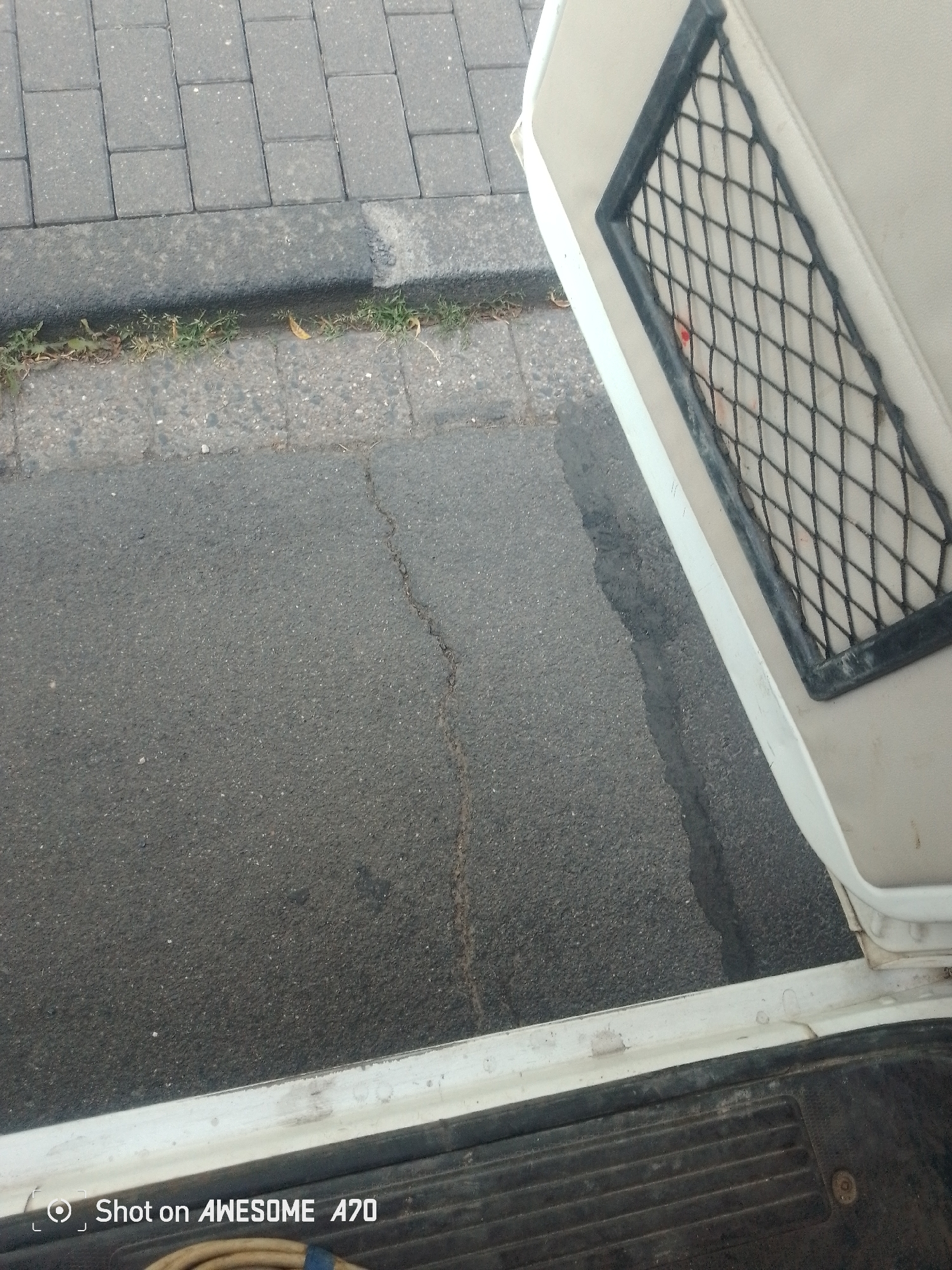 The image shows an outdoor scene with a focus on the edge of a vehicle's interior and the ground outside. In the foreground, the bottom edge of a white vehicle's doorframe is visible, with a black ribbed matting surface on the vehicle's floor. Adjacent to the vehicle is the asphalt of a road, which appears wet in places, suggesting recent rain or damp conditions. A distinct crack runs through the asphalt.
Beyond the road is a grey concrete curb, and behind that, a patterned gray pavement made of interlocking bricks. A thin strip of green grass and some dry leaves grow along the base of the curb.
Within the vehicle's interior, a light-colored seat is visible, with a black mesh-covered compartment on its side. A small red mark is noticeable on the mesh.
The lighting suggests it is daytime, and the presence of wet surfaces implies cloudy or recently rainy weather. The location is identified as Königswinter, Germany. The "Shot on AWESOME A70" text is visible in the bottom left corner, indicating the device used to capture the image. No people or specific events are depicted, making it a static observational shot of a common urban or roadside environment.