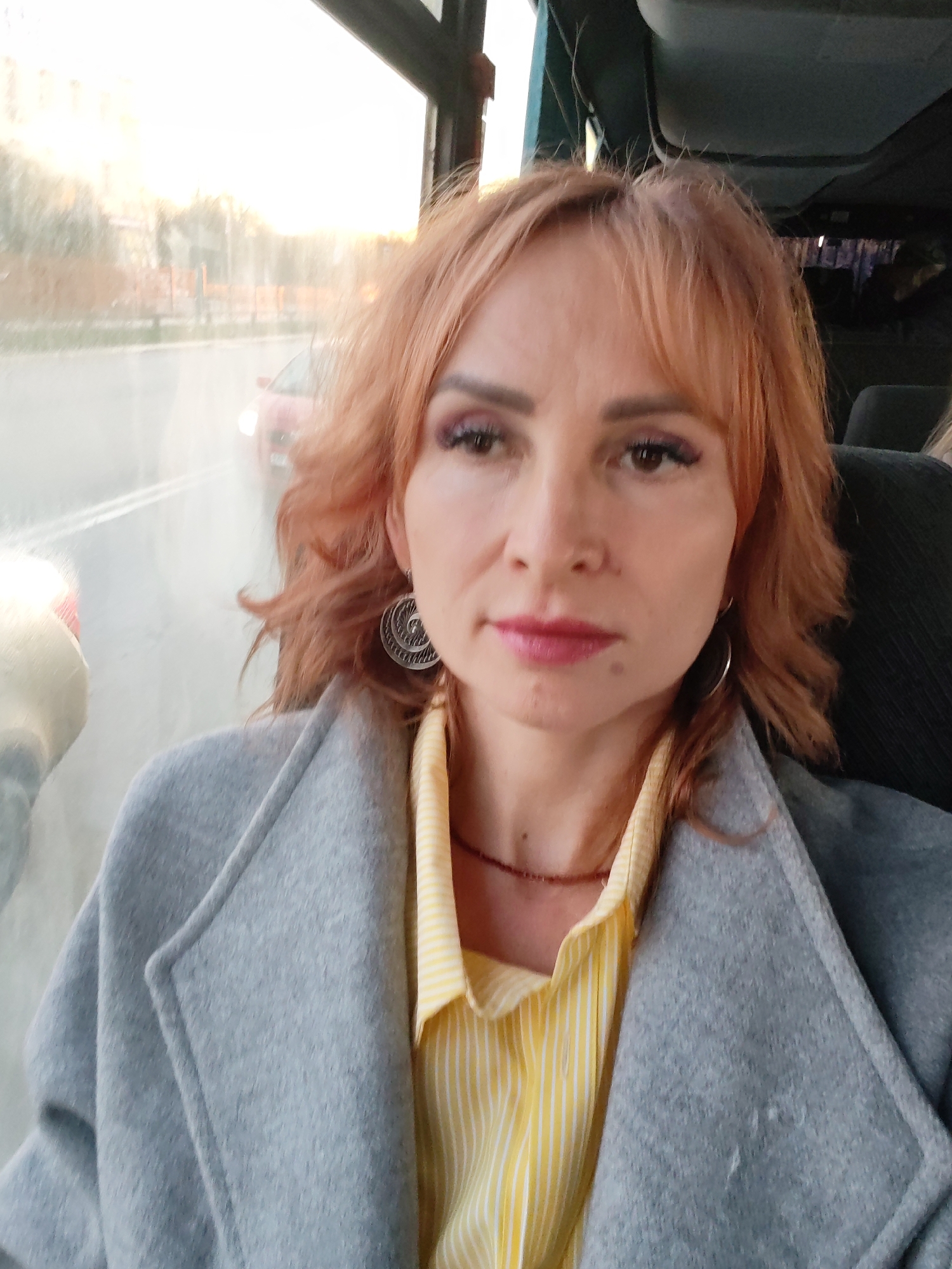 A woman with reddish-blonde hair and fair skin is sitting in what appears to be a vehicle, likely a bus or car, given the seat behind her and the view through the window. She is wearing a light grey wool or felt coat over a yellow and white striped button-down shirt. She is wearing jewelry consisting of silver earrings and a reddish beaded necklace.
Outside the window, there's a road, possibly a city street, with a vehicle visible, suggesting the scene takes place in an urban setting. Based on prior knowledge and the presence of the Chaikovsky identifier, it is likely the picture was taken in Chaikovsky, Russia. There is diffused light streaming in through the window, suggesting either early morning or late afternoon.