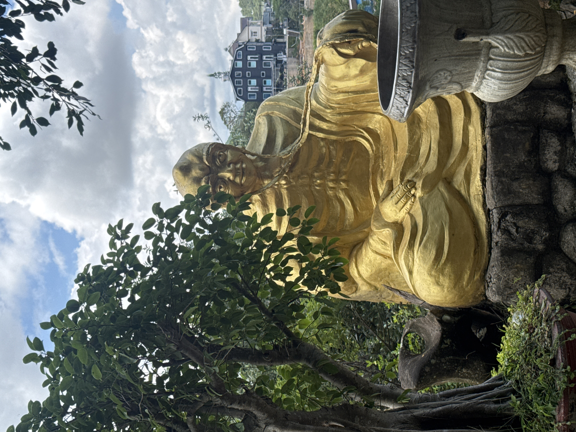 A golden statue of a seated monk is positioned in a lush, green outdoor setting. The statue, which is the primary subject, is life-sized or larger and depicts the monk in traditional robes, with his hands resting in his lap. The statue is made of a material that appears to be gilded or cast in a golden hue, reflecting the sunlight.
To the left of the statue, a large, mature tree with thick branches and abundant green leaves partially obscures the monk. Its trunk is textured and appears to be quite old. A large, dark, abstract-shaped rock formation is situated at the base of the tree and behind the statue's left side. In the foreground, at the lower right, a portion of a decorative, grey stone urn or planter is visible.
In the background, several modern buildings with distinct architectural styles, including multi-story structures with dark facades and prominent windows, are nestled on a hillside. The buildings suggest an urban or semi-urban environment. The sky above is a mix of blue patches and fluffy white clouds, indicating fair weather. The overall scene conveys a sense of tranquility and spirituality, possibly in a temple or garden setting. The location is identified as the Minh Tam Pagoda in Da Lat, Vietnam.
