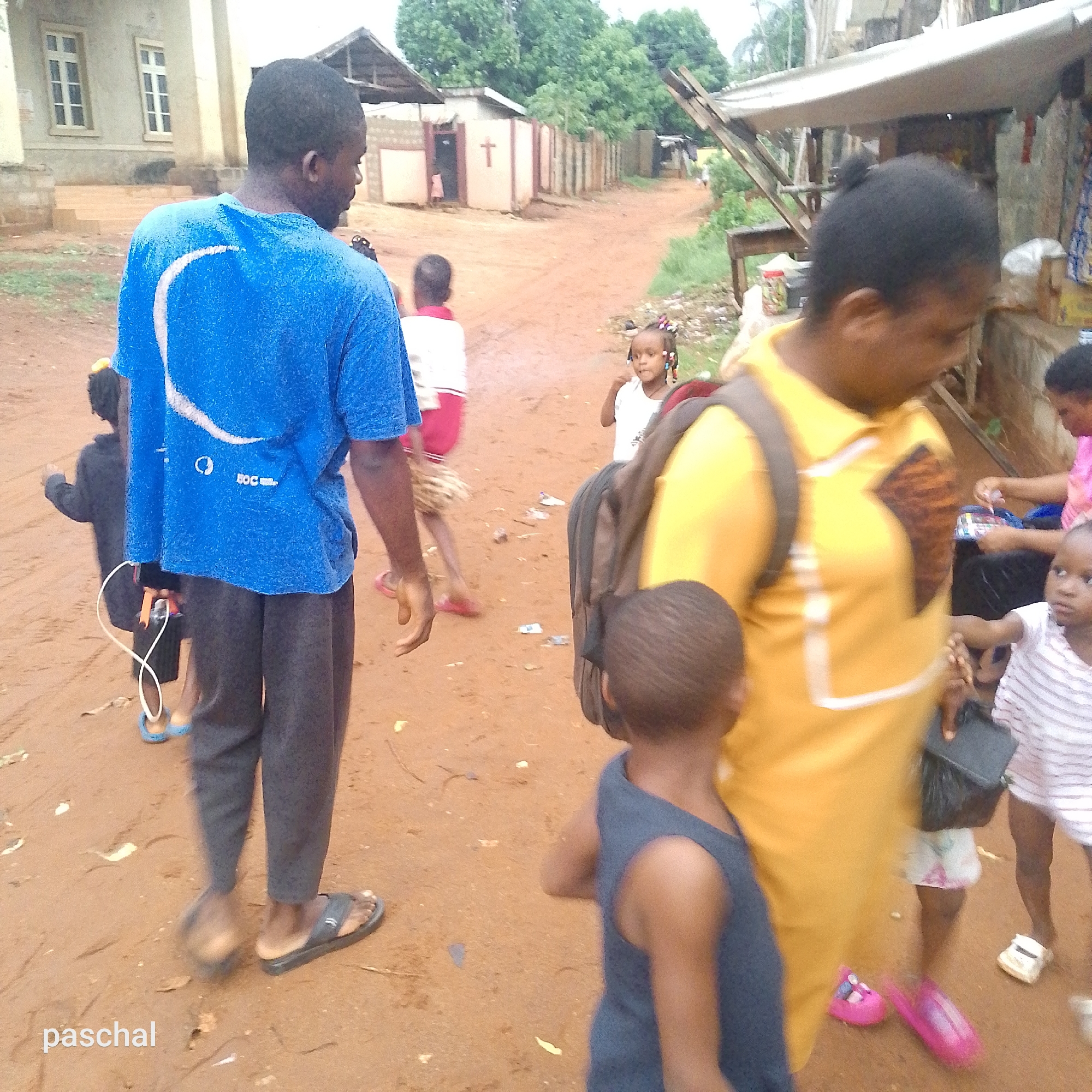 The image depicts a lively street scene on a reddish-brown dirt path, featuring several people, primarily adults and children. In the foreground to the left, a man with dark skin is seen from behind, wearing a blue t-shirt with a white crescent-like design and text that includes "5OC...". He wears dark trousers and flip-flops, holding a white cable or cord. Ahead of him walks a child in a dark outfit.
On the right side of the foreground, a woman with dark skin wears a yellow collared shirt and a backpack, looking downwards. A small child in a blue tank top walks near her. Further right, another woman in a pink top is visible, interacting with a child wearing a striped shirt. Another young child is also present in this group. In the mid-ground, a young girl with braided hair and a white top looks directly at the camera. Another person is seen from behind in the mid-ground left, wearing white and red.
The background reveals a mix of structures and vegetation. To the left, a light-colored building with multiple windows and steps suggests a more permanent structure. In the center, simpler buildings made of concrete blocks line the path; one has a prominent red cross painted on its side, indicating a religious context. On the far right, a makeshift stall with a white tarp roof is visible, suggesting a market or shop. The lighting indicates it is daytime, possibly overcast. The word "paschal" is watermarked in the bottom left corner.