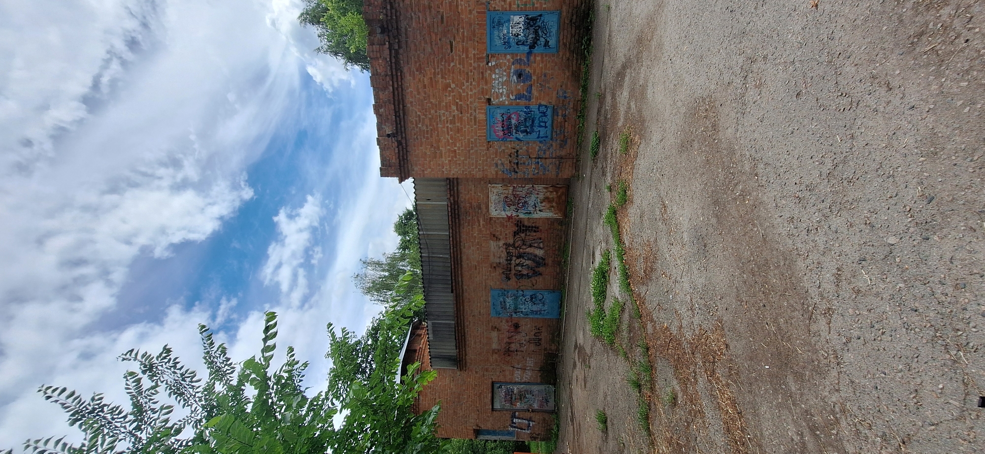 This image, taken in Bila Tserkva, Ukraine, depicts an exterior view of a weathered, brick building adorned with graffiti. The building appears to be in a state of disrepair, with several framed openings on its brick facade covered with colorful, abstract spray paint. Some of these frames are blue, while others are a rusty metal. The brickwork itself shows signs of age and wear.
Adjacent to the building is a paved area, possibly a road or courtyard, with a rough, granular surface. Scattered patches of green grass and dry, brown debris are visible on this surface, suggesting it's an outdoor, possibly neglected, area. Lush green trees and foliage are visible in the background and to the left of the building, partially obscuring the sky.
The sky above is a prominent feature, filled with dramatic, wispy white clouds against a bright blue backdrop. The lighting suggests it is daytime, likely late morning or early afternoon, given the clarity and brightness of the sky. There is no discernible human activity in the frame. The overall impression is one of an urban or semi-urban environment, characterized by the contrast between the decaying building and the vibrant natural elements of the sky and trees. The graffiti indicates a potential artistic or subcultural presence in the location. No readable text is visible on the graffiti or elsewhere in the image.