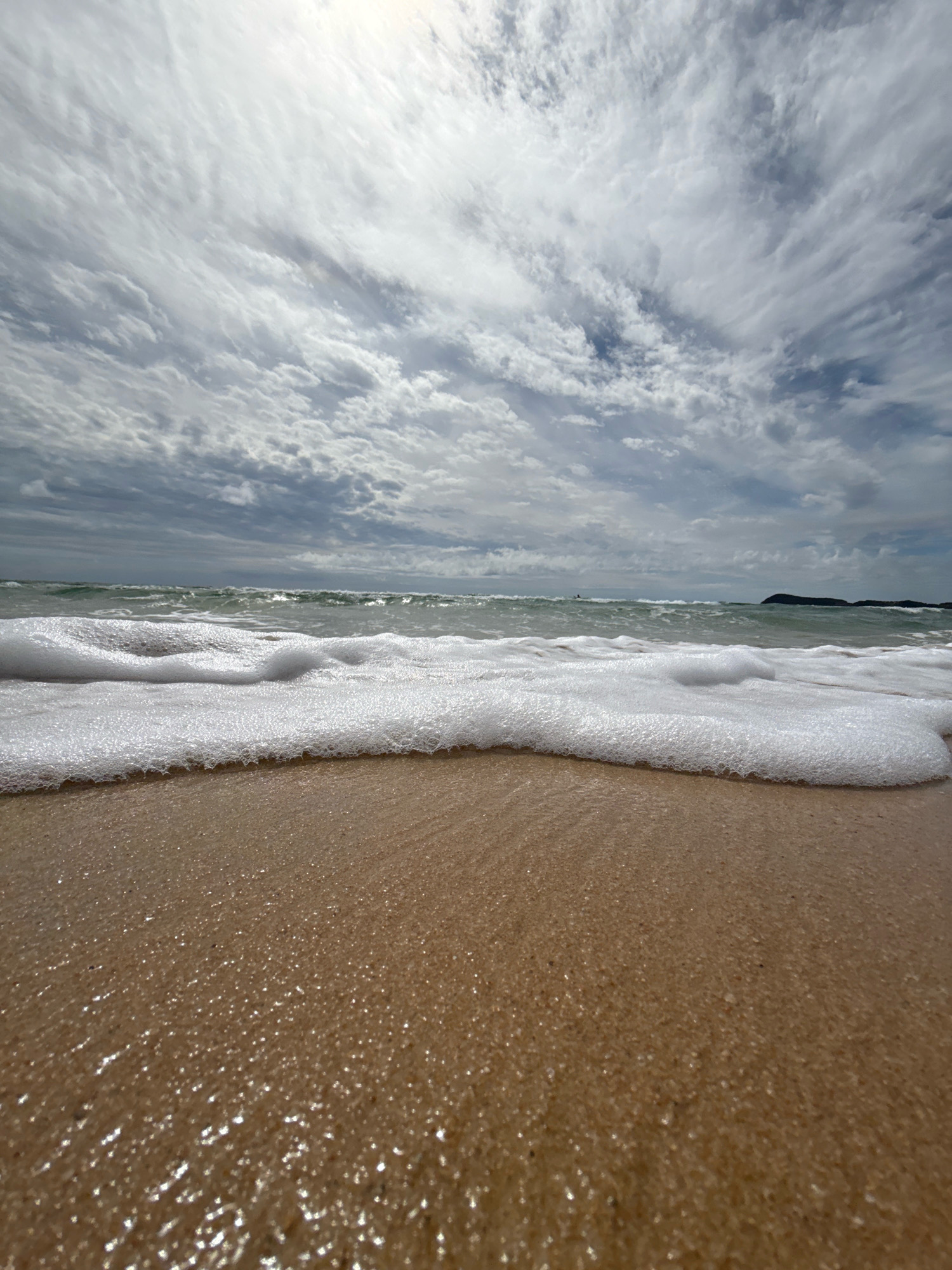The image depicts a low-angle view of a beach with waves lapping at the shore. The foreground is dominated by wet sand, reflecting the light. A line of white sea foam extends across the middle ground, marking the edge of the incoming tide. Beyond the foam, the ocean stretches out, with gentle waves and a hazy horizon. The sky is filled with dramatic, wispy clouds, suggesting an overcast day with patches of sunlight breaking through. The scene is devoid of any people or man-made structures, creating a natural and tranquil atmosphere. The location is identified as the city of Anna, Russia.