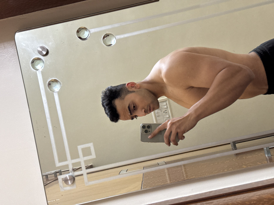 A shirtless man stands in front of a mirror, holding a phone in his right hand to take a selfie. He wears dark shorts. The mirror has a design with geometric shapes, including circles and lines, and it reflects a wall with a light switch. The mirror frame is partially visible at the top and bottom of the image.