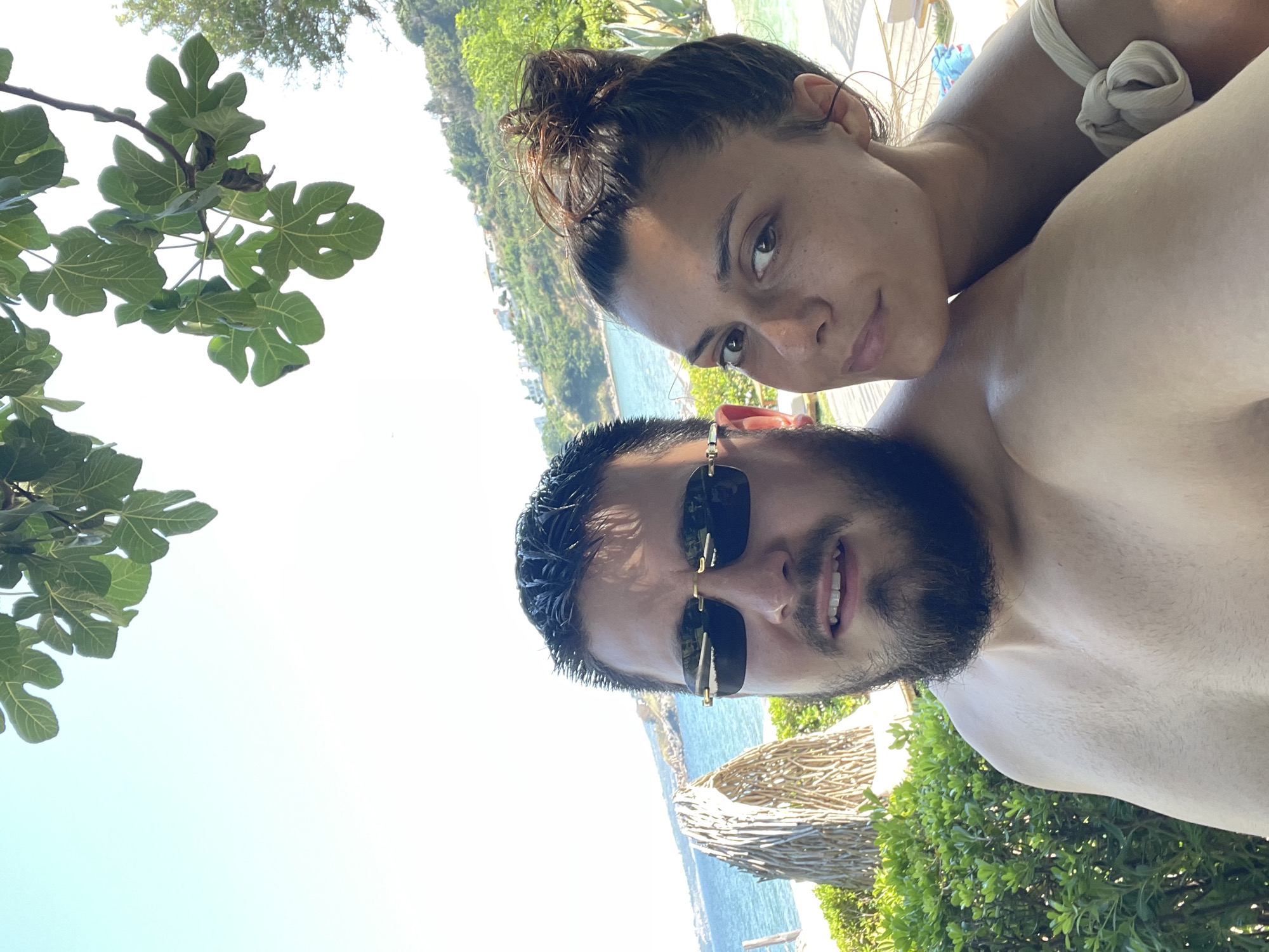 A selfie captures a couple posing outdoors on a sunny day, with a beach and coastal landscape in the background. The man, in the foreground, wears sunglasses and has a beard. The woman is positioned behind him, leaning her head on his shoulder. Lush green foliage and a structure made of driftwood are visible behind them. The sea stretches out to the horizon, with buildings dotting a hillside in the distance. The sky is clear and bright. The location is implied to be Chernomorets, Bulgaria.