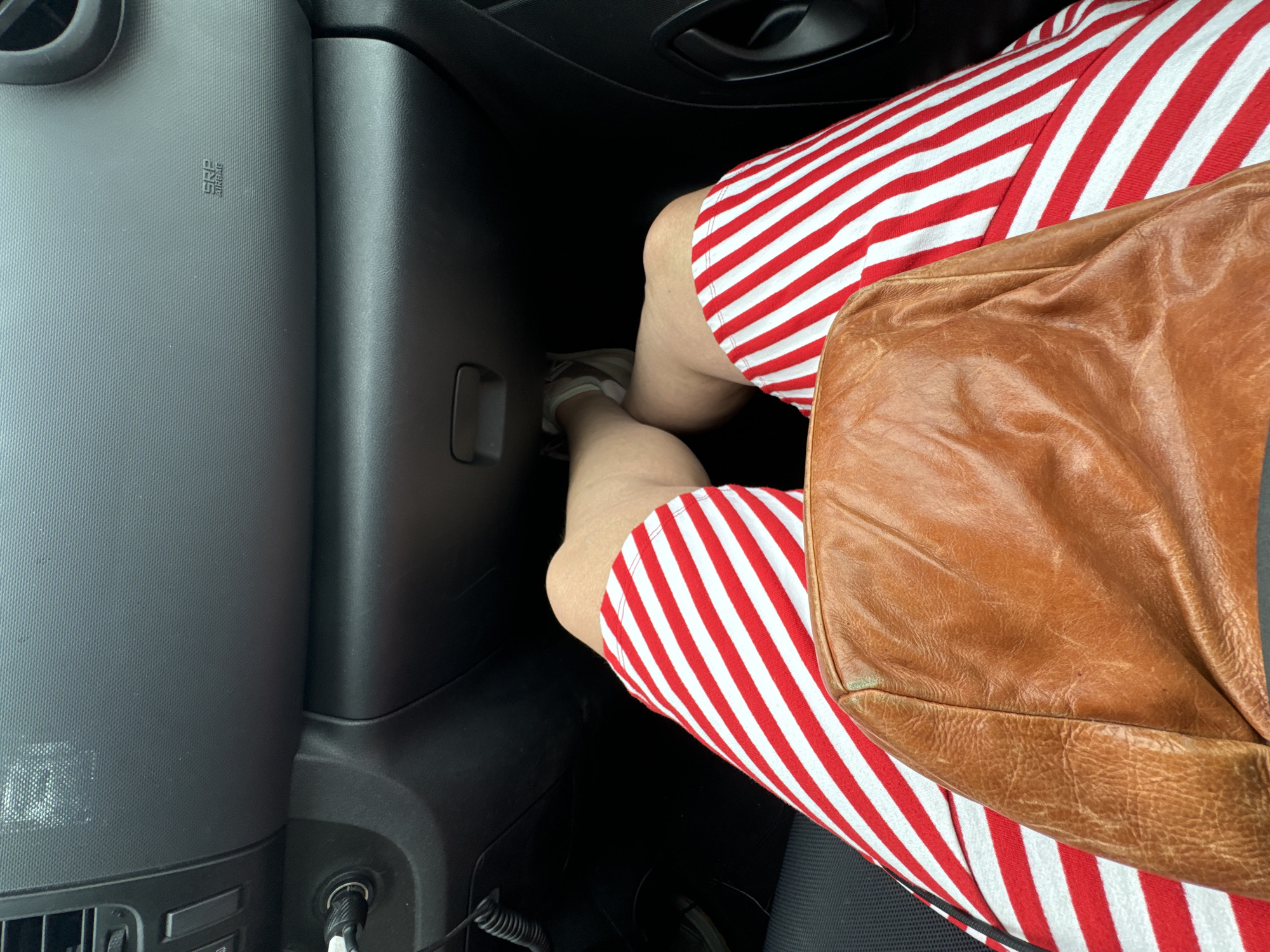 The image shows a person's legs and lap inside a car, with a brown leather bag resting on their lap. The person is wearing red and white striped shorts. The car's dashboard is visible in the background, with an "SRS AIRBAG" label on it. The scene is captured from a high-angle perspective, looking down at the person's legs. The location is specified as Kilcurl, Ireland.