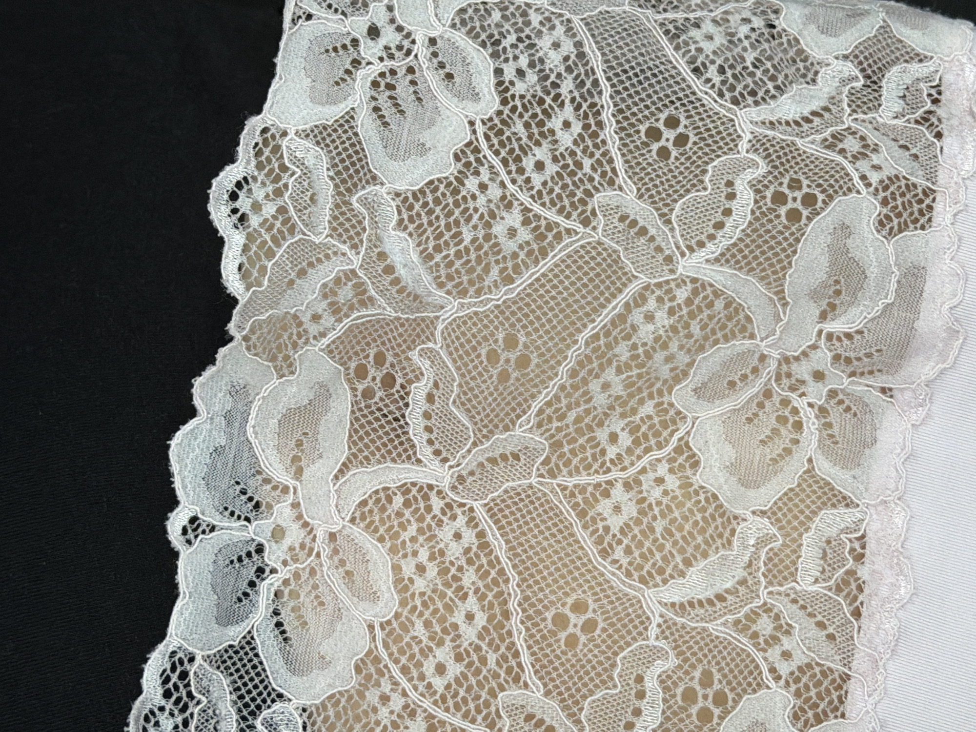 A close-up, overhead shot shows a delicate white lace trim against a dark background. The lace features a floral pattern with intricate interwoven threads creating an open, net-like texture. The top edge of the lace is attached to a slightly lighter fabric. The bottom edge of the lace is scalloped. The image does not contain any identifiable elements that would allow for its location in the city of Chaikovsky, Russia, or any other specific location.