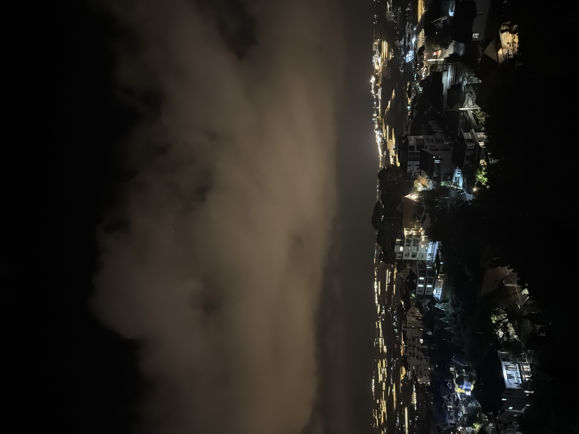 A nighttime aerial view captures a cityscape in Xuân Trường - Đà Lạt, Vietnam, under a cloudy sky. Numerous buildings, illuminated by interior lights and streetlights, dot the landscape. In the foreground, closer structures are visible, some with multiple stories and windows casting warm light. Trees and foliage are scattered throughout, appearing as dark silhouettes against the lit areas. In the distance, the city stretches out with a dense concentration of illuminated buildings and what appear to be greenhouses or agricultural structures, their lights forming parallel rows. The clouds in the sky are illuminated from below by the city lights, giving them a soft, diffused glow. The overall impression is of a vibrant, living city at night, partially obscured by atmospheric conditions.