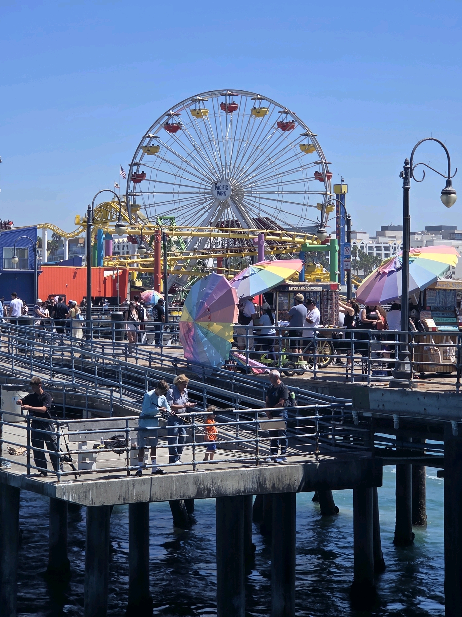 This is a photograph of the Santa Monica Pier in Santa Monica, California, United States of America, on a clear, sunny day. In the background, a large Ferris wheel with red and yellow gondolas is visible, along with the tracks of a roller coaster. The pier itself is bustling with activity. Several people are walking along the wooden walkway, some are browsing at vendor stalls with colorful umbrellas selling kites and souvenirs. In the foreground, on a lower level of the pier, a group of people are fishing off the edge. A young boy in an orange shirt is among them, along with adults holding fishing rods. The water below the pier is a deep blue, with ripples and reflections. The scene captures a lively atmosphere characteristic of a popular tourist destination.