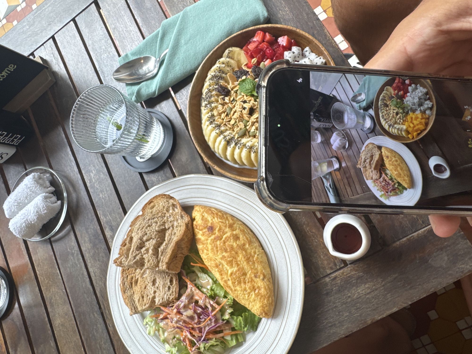 The image depicts a dining table set with several dishes. A smoothie bowl with bananas, granola, strawberries, and dragon fruit sits in a wooden bowl. A plate of bread, an omelet, and salad are on a white plate. A glass of water sits on a coaster, and a small bowl with a dark sauce is also present. A smartphone is held over the table, capturing a photo of the food. The scene is set in what appears to be an outdoor dining area in Đà Nẵng, Vietnam.