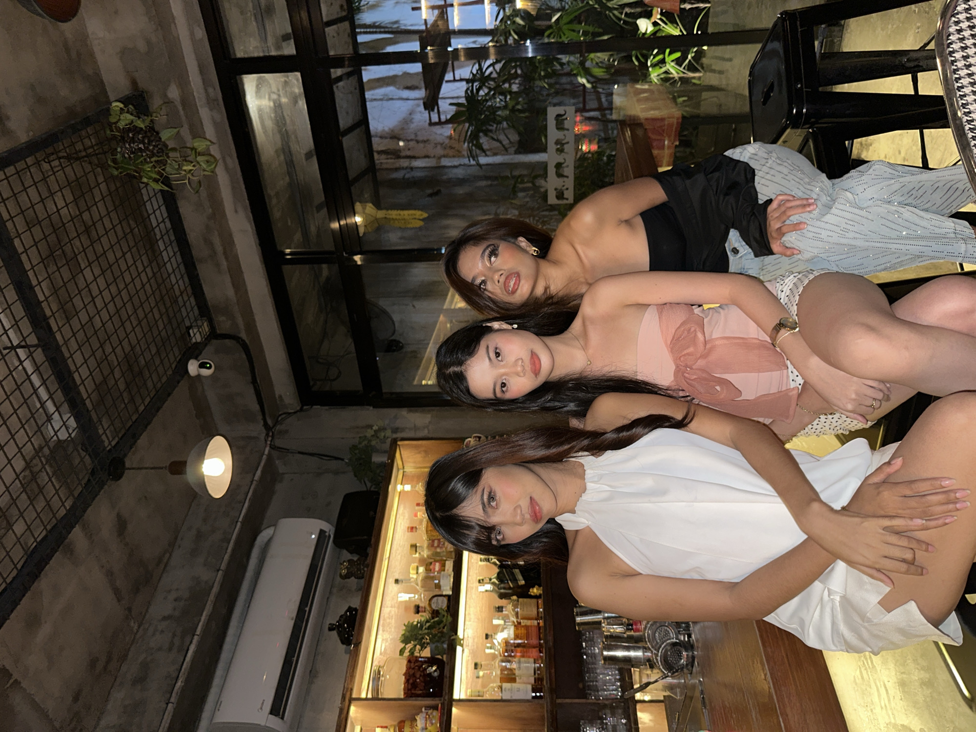 Three young women are seated at a bar in what appears to be a dimly lit establishment in Davao City, Philippines. The woman on the left is wearing a white halter dress and has long, dark hair. She is looking directly at the camera with a neutral expression. The woman in the middle is wearing a pink strapless top with a large bow and a white skirt with polka dots. Her hair is dark and she is also looking forward. The woman on the right is wearing a black strapless top and light-colored pants adorned with rhinestones. She has dark hair and is looking slightly to her left, with a playful expression. Behind them, shelves are stocked with bottles of alcohol and other bar supplies. The interior features exposed concrete walls and ceiling, industrial lighting, and potted plants, suggesting a modern or trendy bar setting.